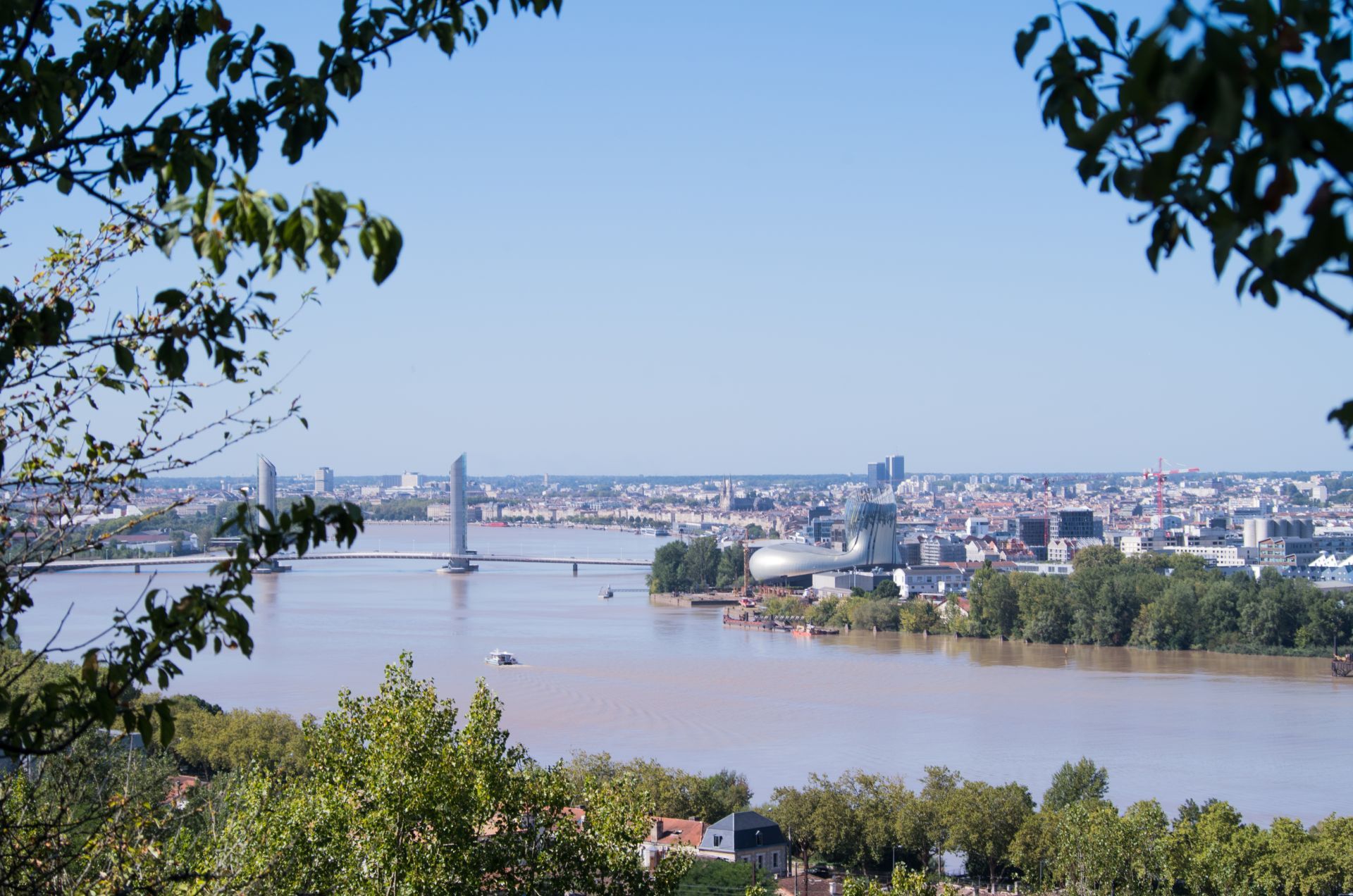 Vue panoramique de Bordeaux avec le pont de Pierre enjambant la Garonne, la cathédrale et les bâtiments historiques de la ville en arrière-plan, encadrés par la végétation.