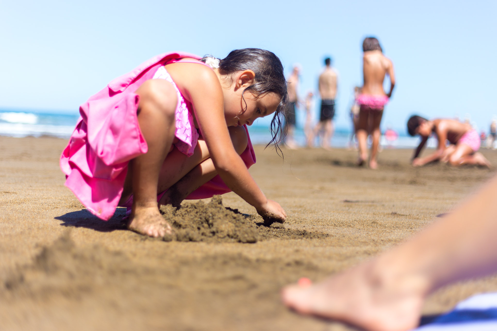 Une enfant en maillot de bain rose joue dans le sable sur une plage, avec d'autres baigneurs en arrière-plan et l'océan sous un ciel dégagé.
