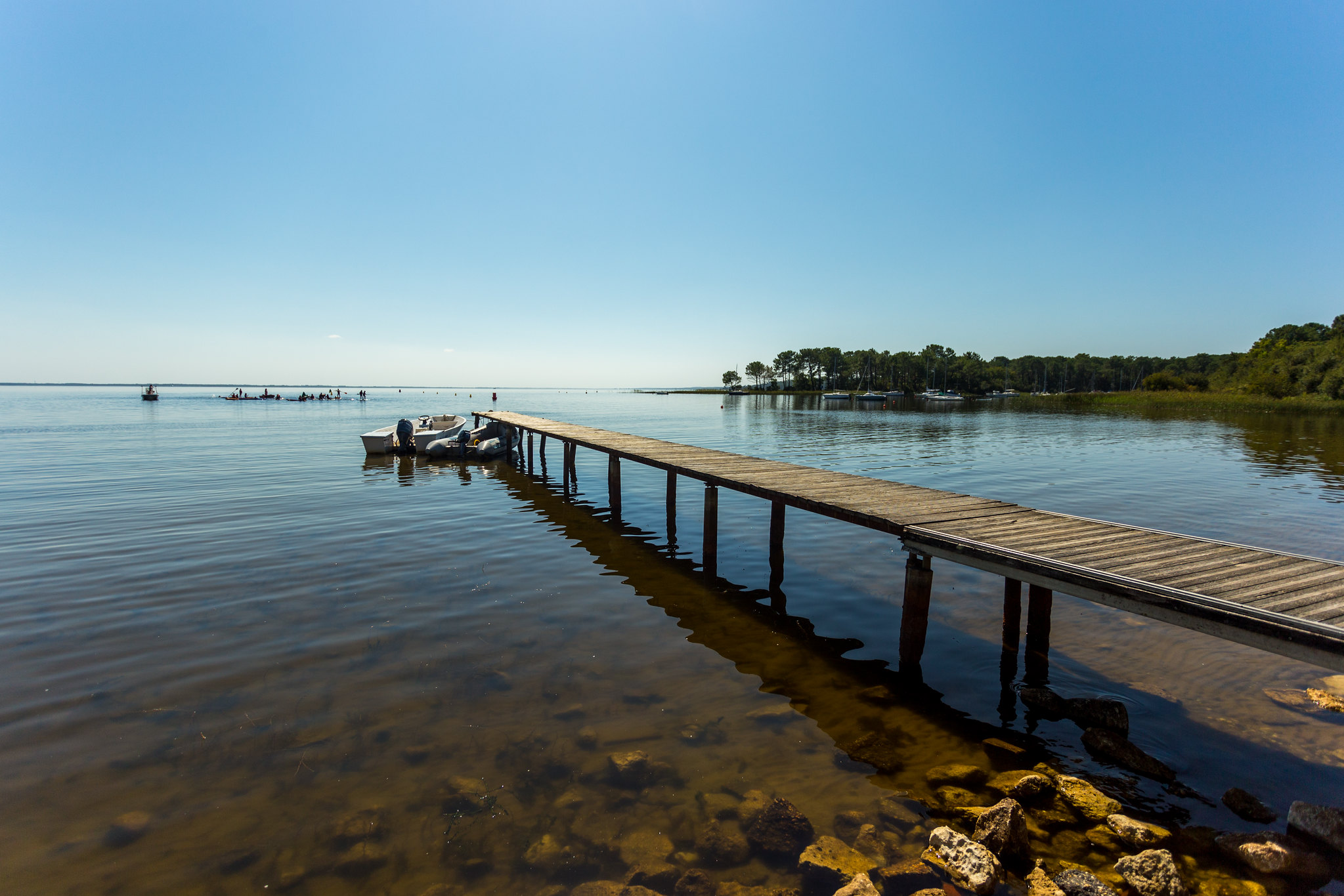 Un long ponton en bois s'étend dans une baie calme avec un petit bateau amarré, des arbres en arrière-plan et un ciel bleu clair, paysage fluvial typique de la région de Bordeaux.