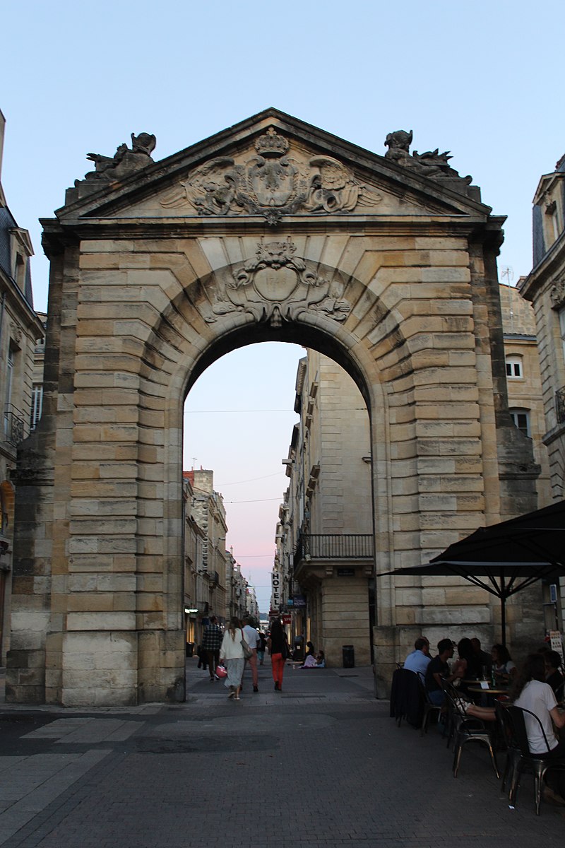 Porte de la Grosse Cloche à Bordeaux, imposant arc de pierre de style Renaissance avec bas-reliefs, donnant sur une rue pavée avec façades haussmanniennes et terrasse de café en premier plan.