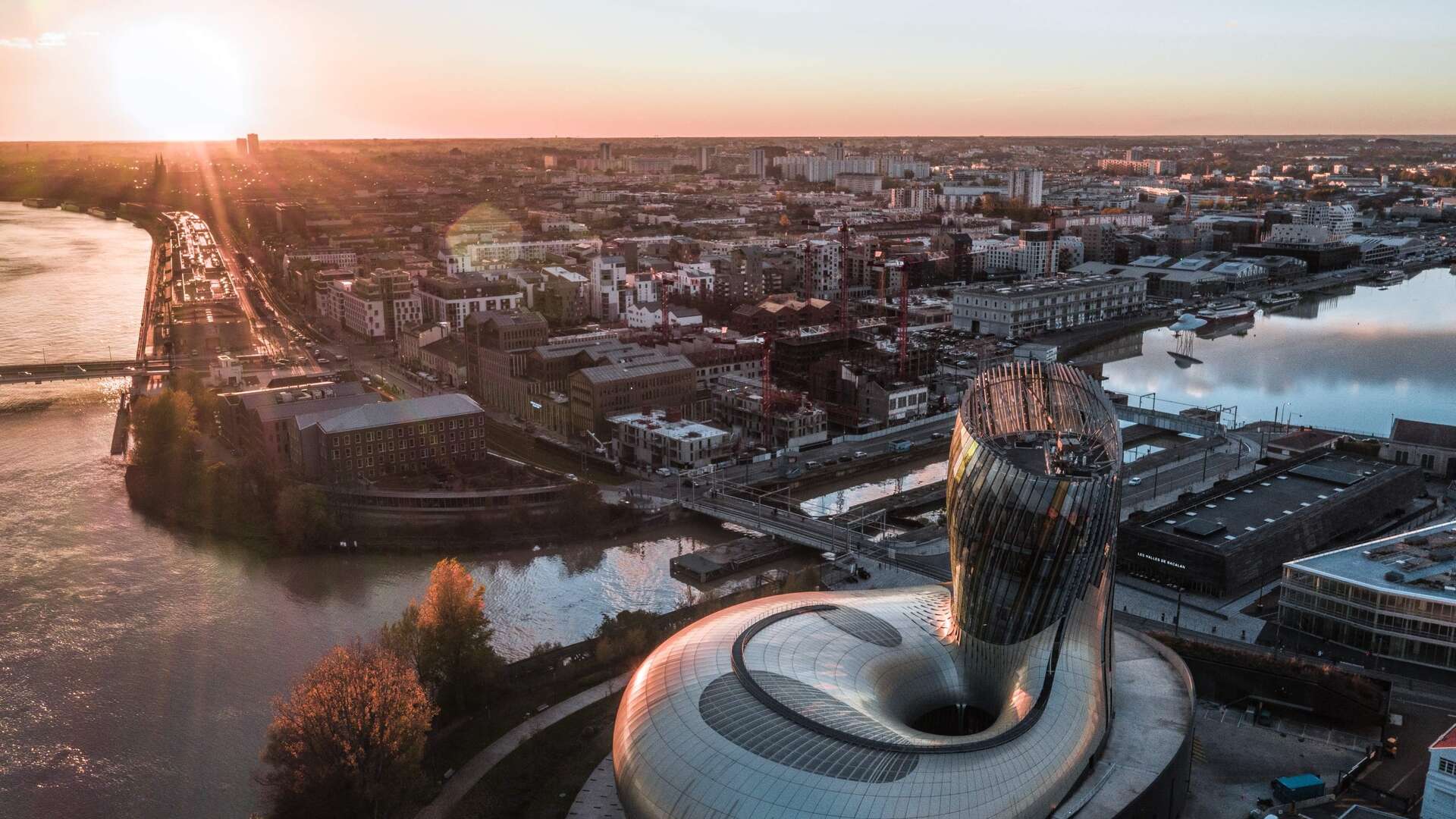 Vue aérienne au coucher de soleil des Bassins à flot de Bordeaux avec l'architecture moderne du Muséum d'Histoire Naturelle au premier plan, la Garonne et la ville en arrière-plan.