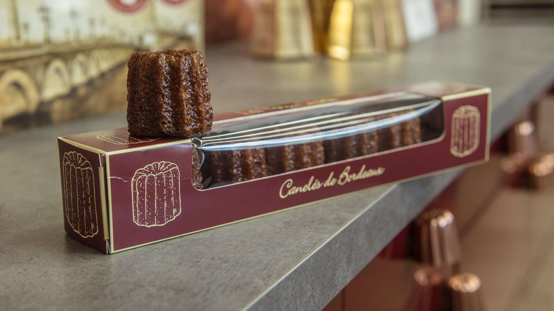 Canelé de Bordeaux dans sa boîte d'emballage traditionnelle marron et or, posé sur une surface grise avec d'autres canelés en arrière-plan