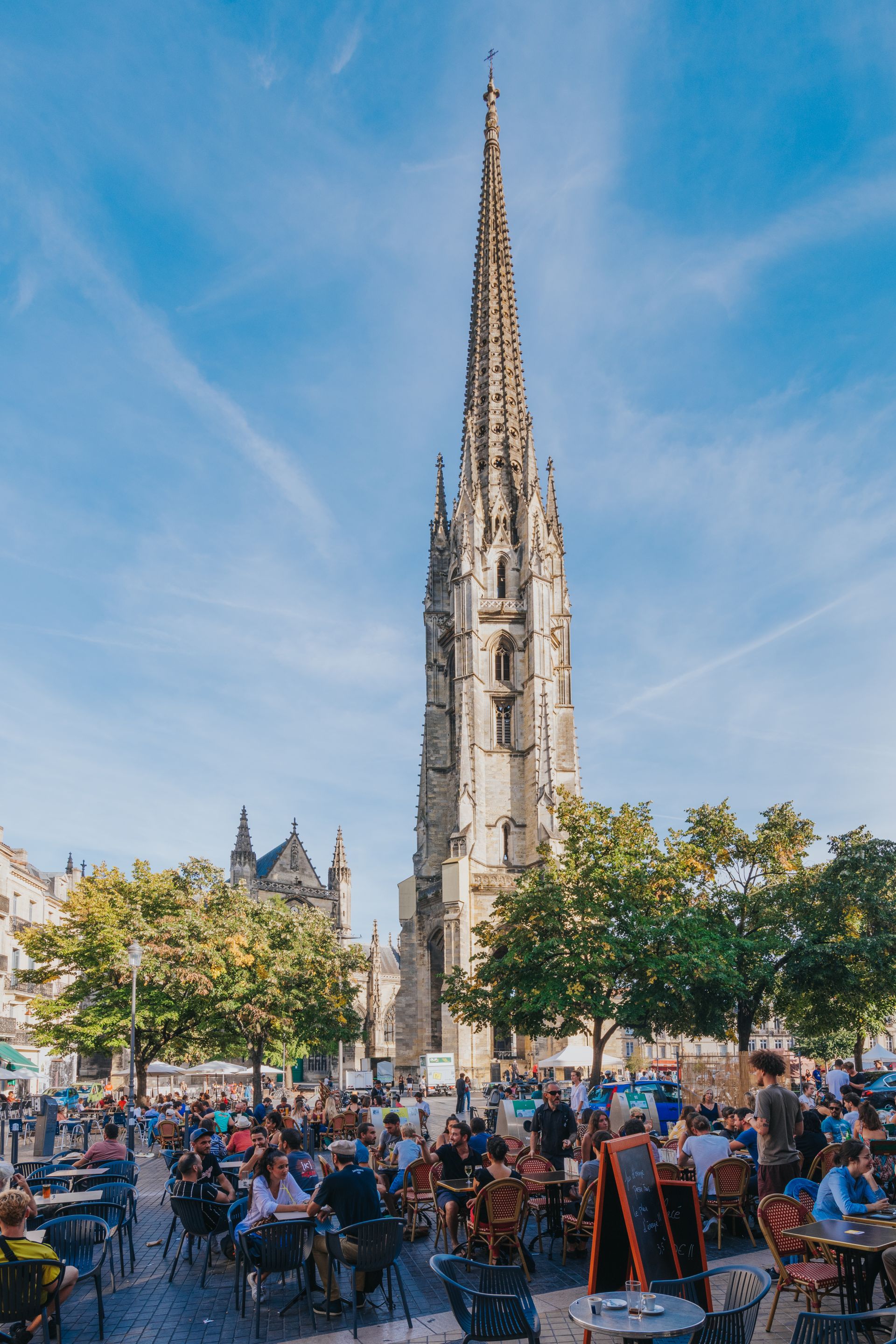 La Basilique Saint-Michel de Bordeaux avec sa flèche gothique élancée dominant une place animée remplie de terrasses de cafés et de visiteurs sous un ciel bleu.