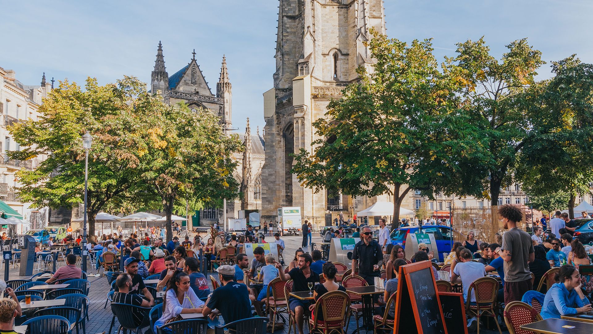 Vue d'une terrasse animée sur une place de Bordeaux avec la basilique Saint-Michel en arrière-plan, sous un ciel bleu dégagé.