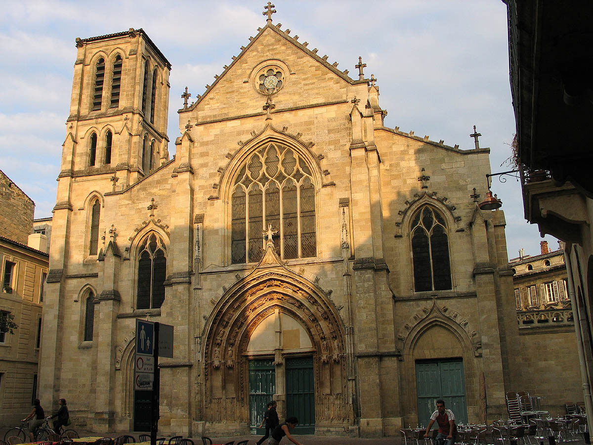 Église gothique de Bordeaux avec façade en pierre blonde, tour carrée, grandes baies vitrées et portail en arche ogivale, entourée de bâtiments historiques.