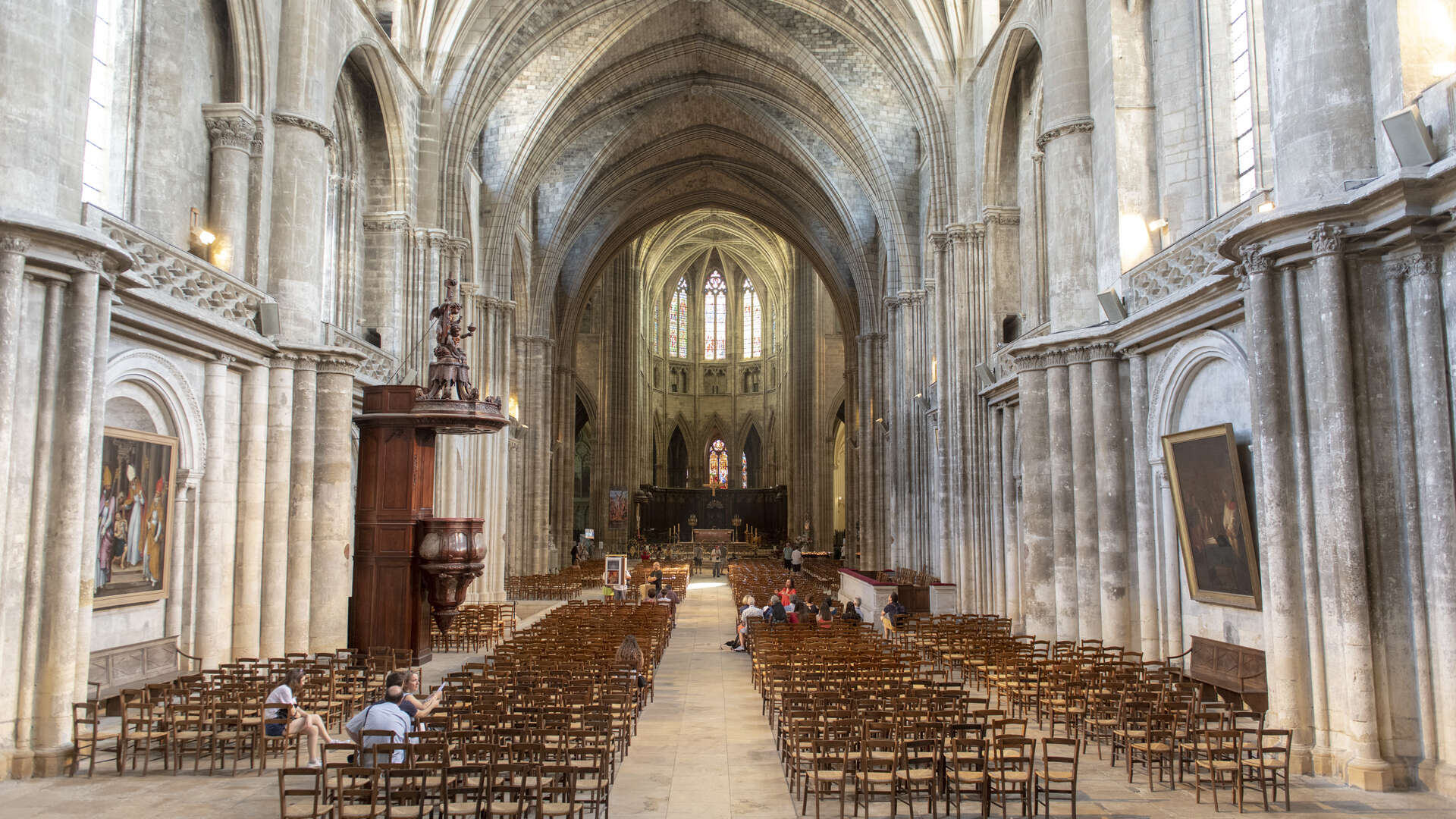 Intérieur de la Cathédrale Saint-André de Bordeaux montrant la nef centrale avec ses voûtes gothiques, ses colonnes en pierre, ses bancs en bois et l'autel illuminé au loin.