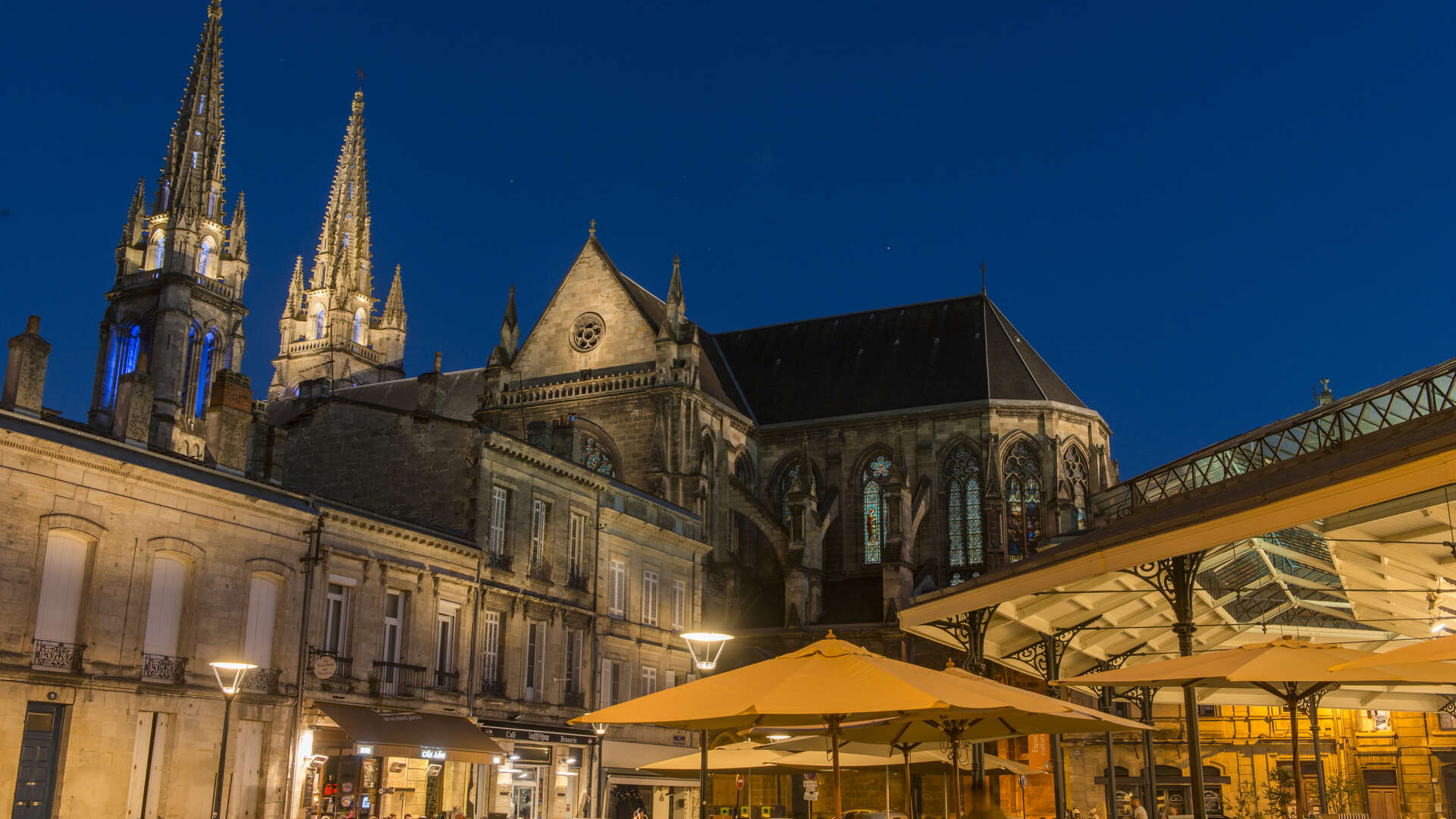 Vue nocturne de la Basilique Saint-Michel de Bordeaux avec ses flèches gothiques illuminées, entourée de bâtiments historiques et de terrasses ombragées en premier plan.