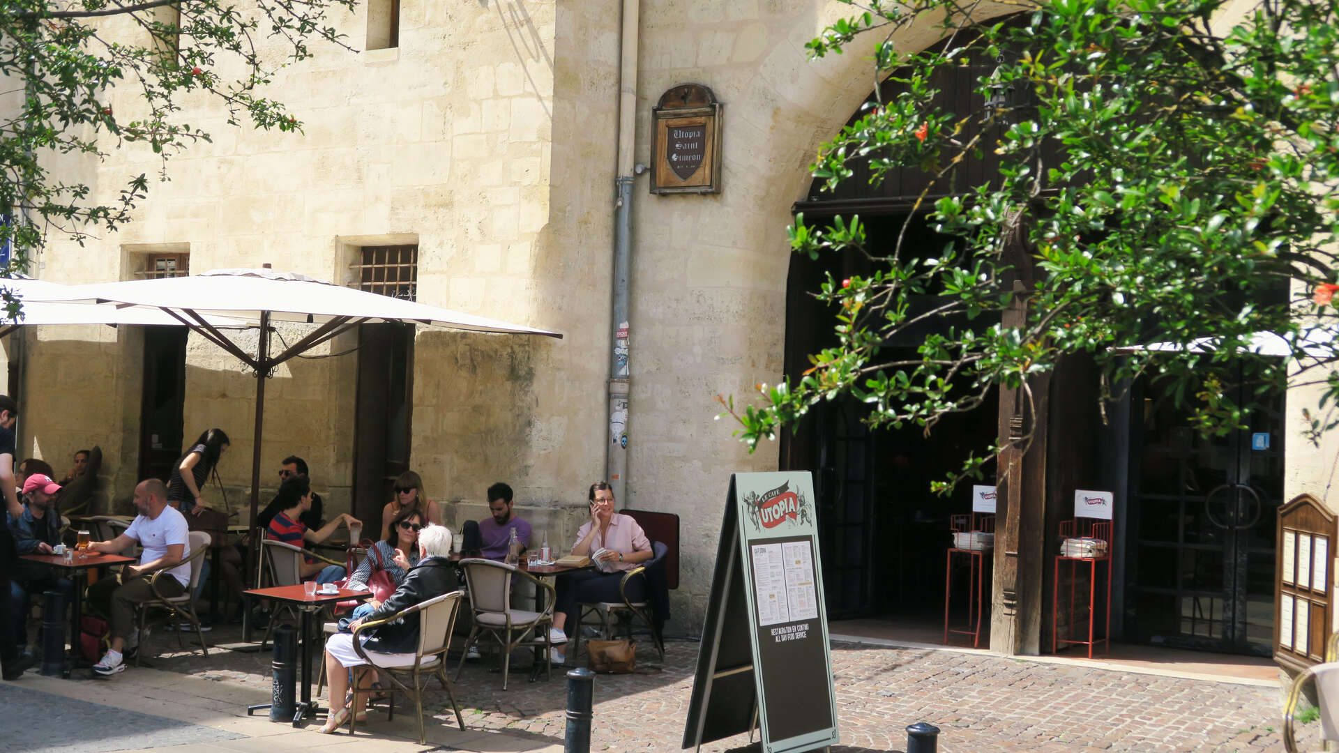 Terrasse extérieure du Cinéma Utopia à Bordeaux avec clients assis sous un parasol blanc, façade en pierre et végétation grimpante.
