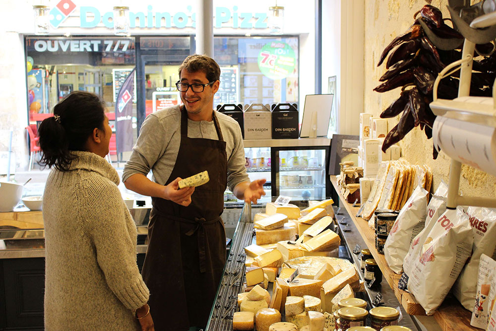 Un fromager sourit en montrant du fromage à une cliente dans sa boutique bordelaise. Le comptoir est rempli de fromages français variés et de produits gastronomiques.