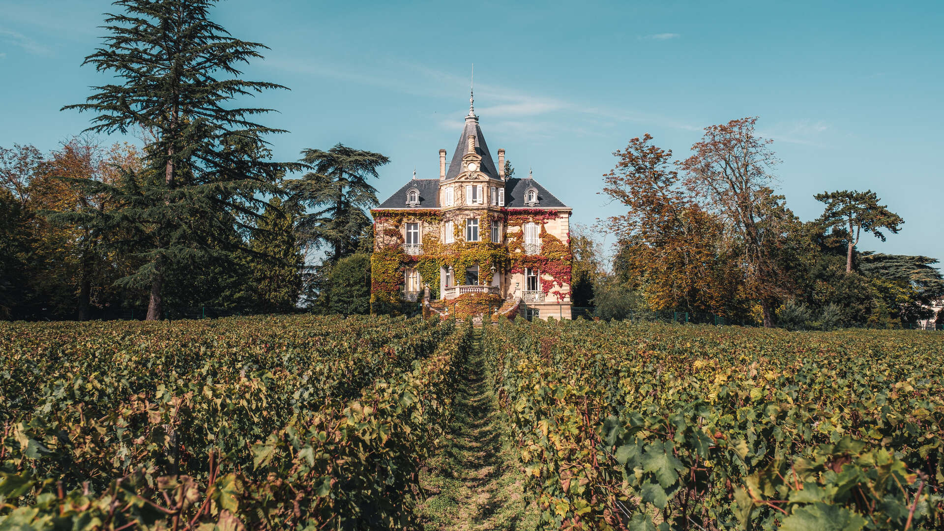 Château viticole en briques rouges avec tourelle et toit pointu, entouré de vignobles en automne près de Bordeaux