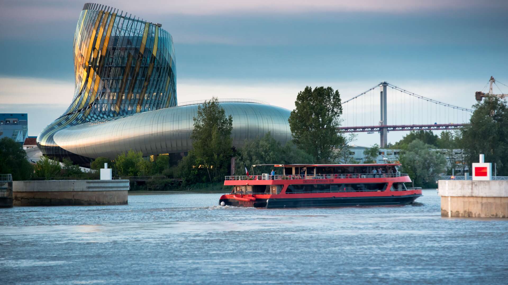 Un bateau de croisière rouge navigue sur la Garonne devant la Cité du Vin et le pont de Pierre à Bordeaux, avec l'architecture moderne du musée en arrière-plan