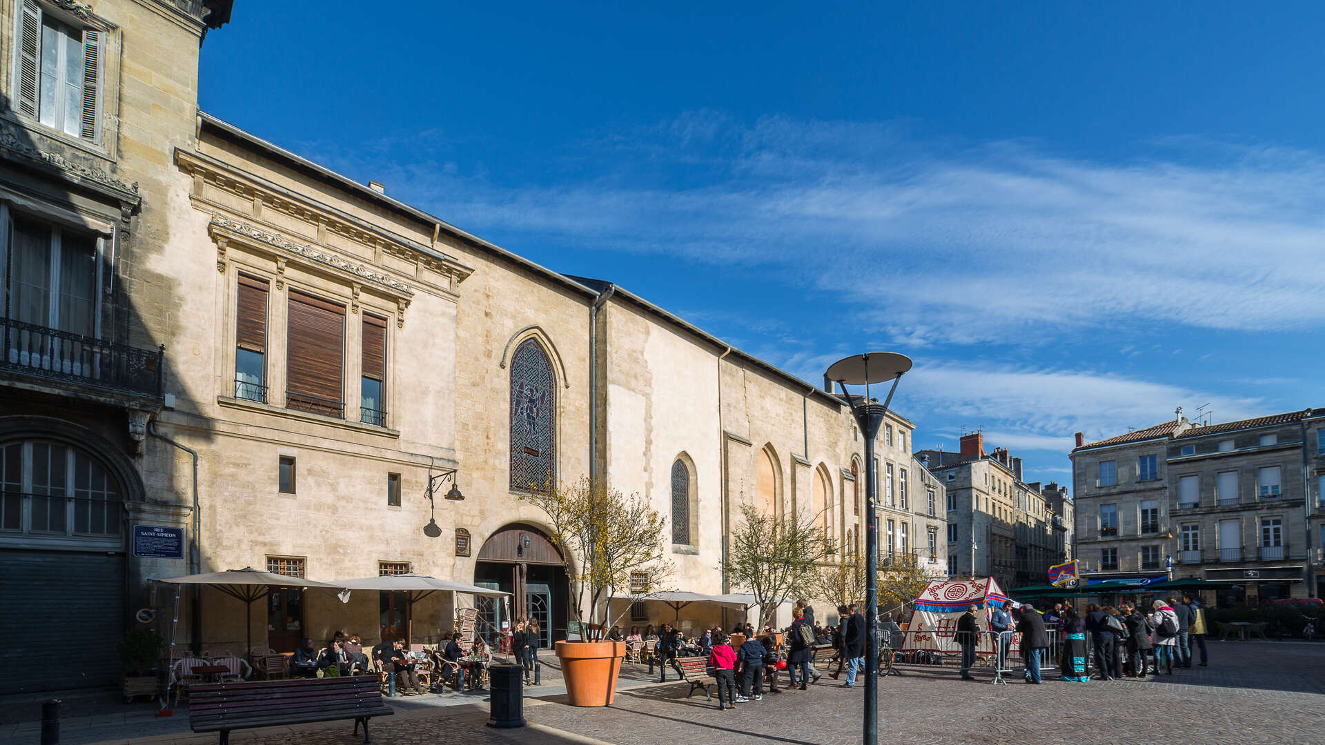 Façade du Cinéma Utopia à Bordeaux, bâtiment historique en pierre avec arcade, terrasse avec parasols, place publique animée avec visiteurs et bâtiments haussmanniens en arrière-plan.