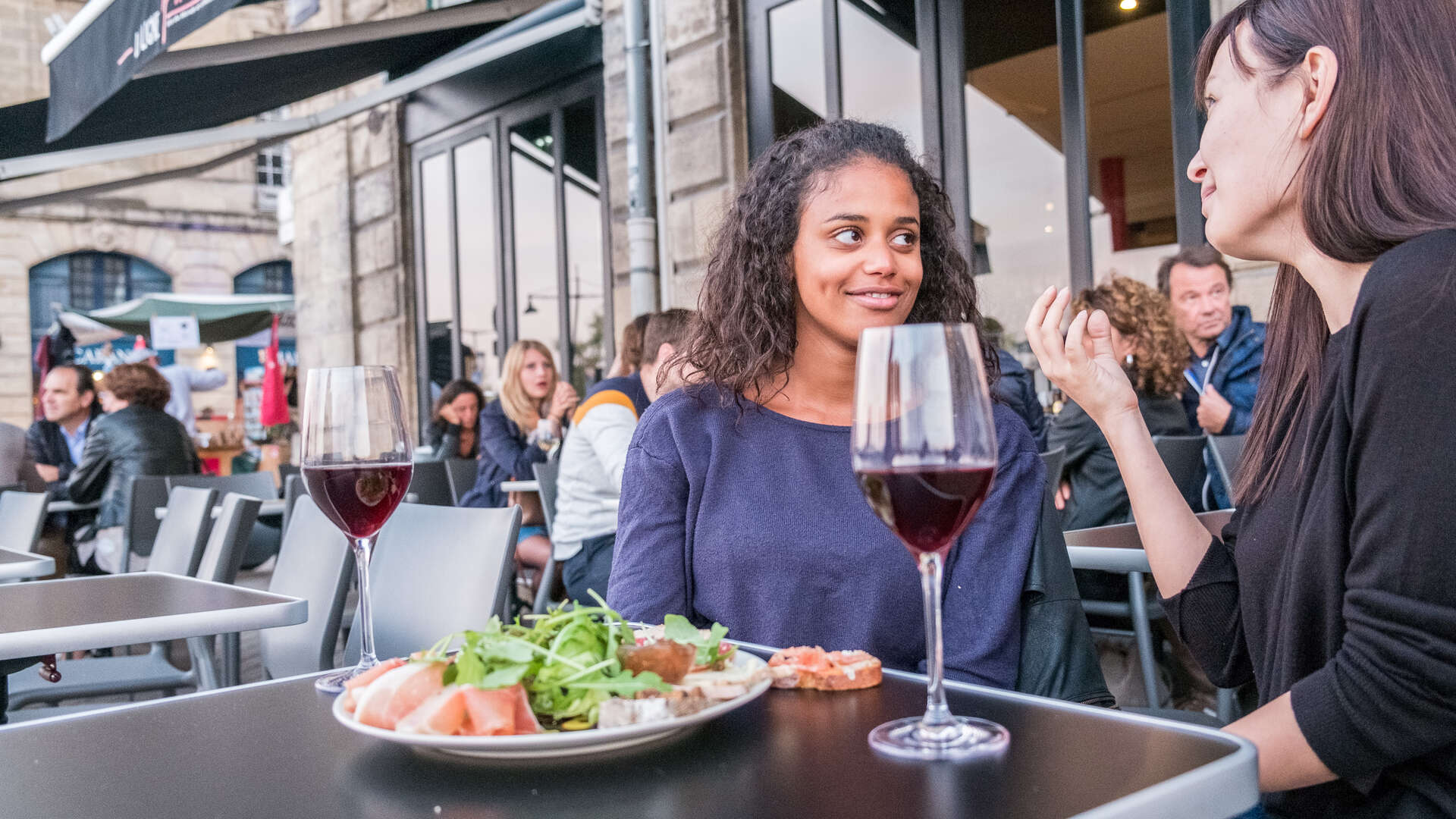 Deux femmes dégustent du vin rouge et un plat de fruits de mer en terrasse à Bordeaux, entourées d'autres clients dans un restaurant urbain.
