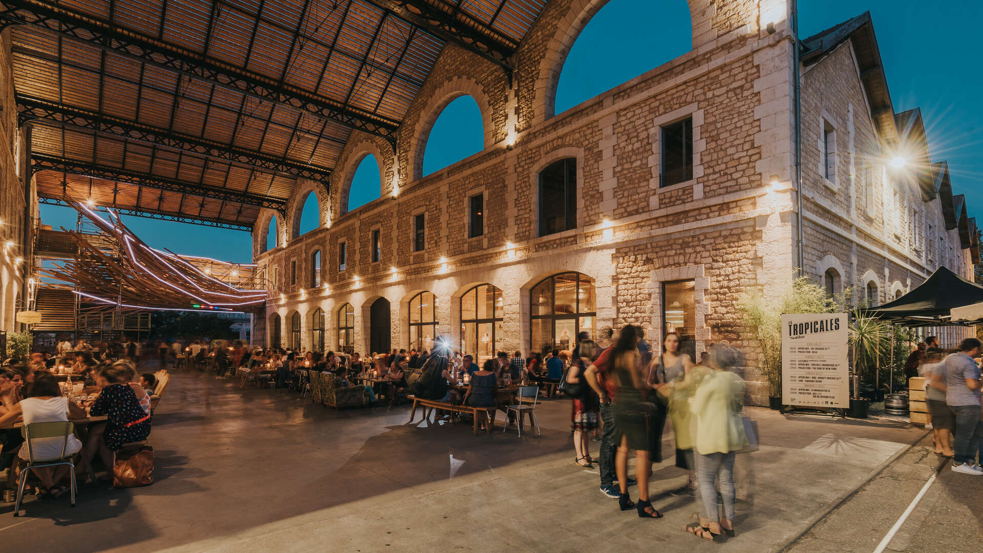 Soirée animée dans la cour intérieure de Darwin écosystème à Bordeaux, avec des visiteurs assis autour de tables, sous une structure métallique couverte et des arcades en pierre illuminées au coucher du soleil.