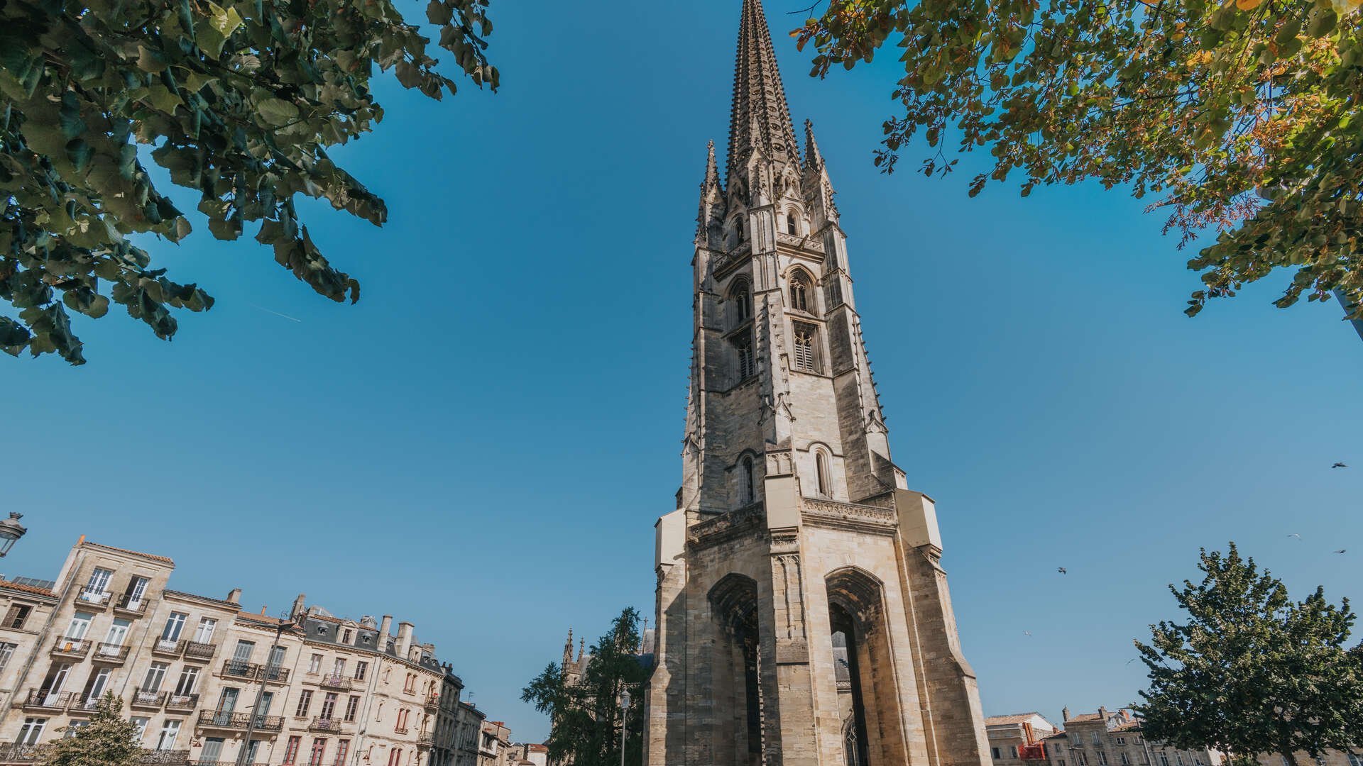 La Flèche Saint-Michel de Bordeaux, église gothique avec son clocher élancé, photographiée depuis une place entourée de bâtiments haussmanniens sous un ciel bleu dégagé.