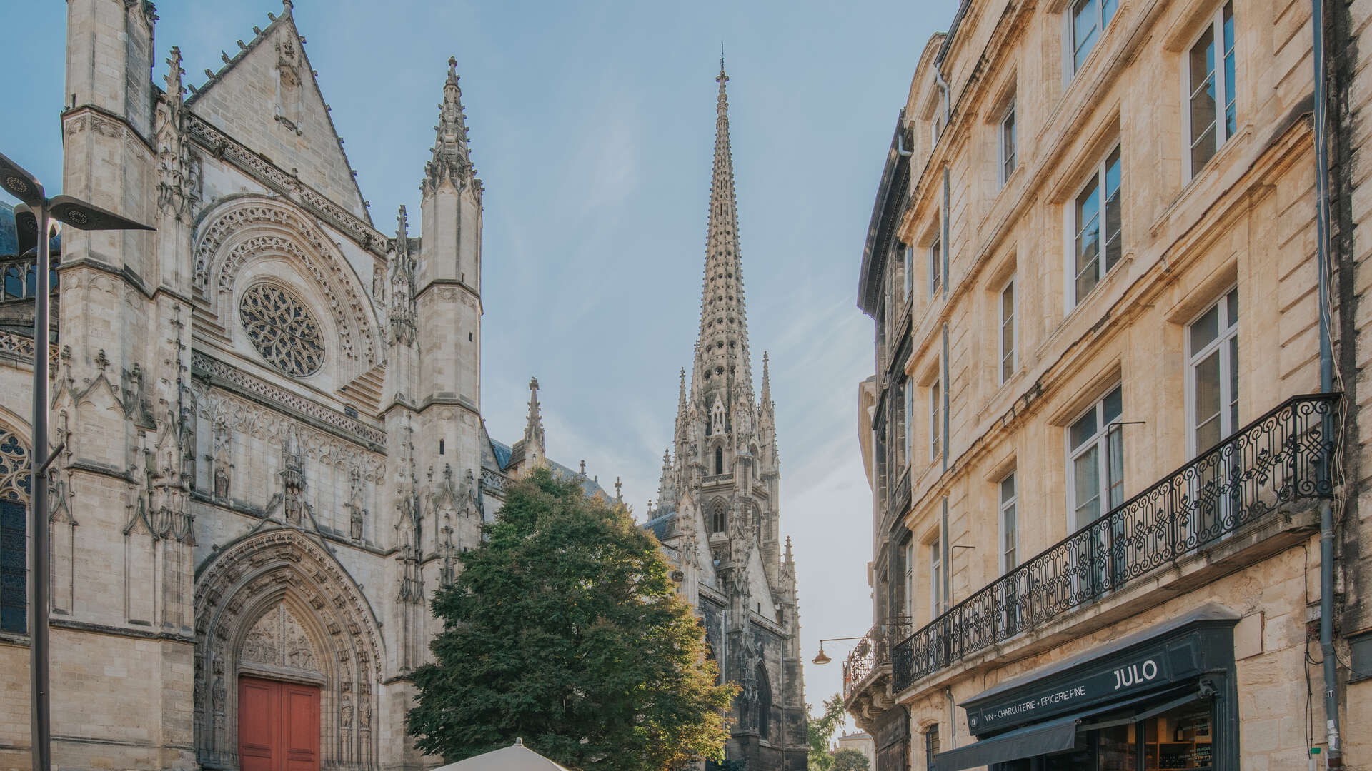 Vue de la flèche Saint-Michel de Bordeaux, église gothique avec sa tour pointue caractéristique, encadrée par des bâtiments haussmanniens et une boutique JULO en bas à droite.
