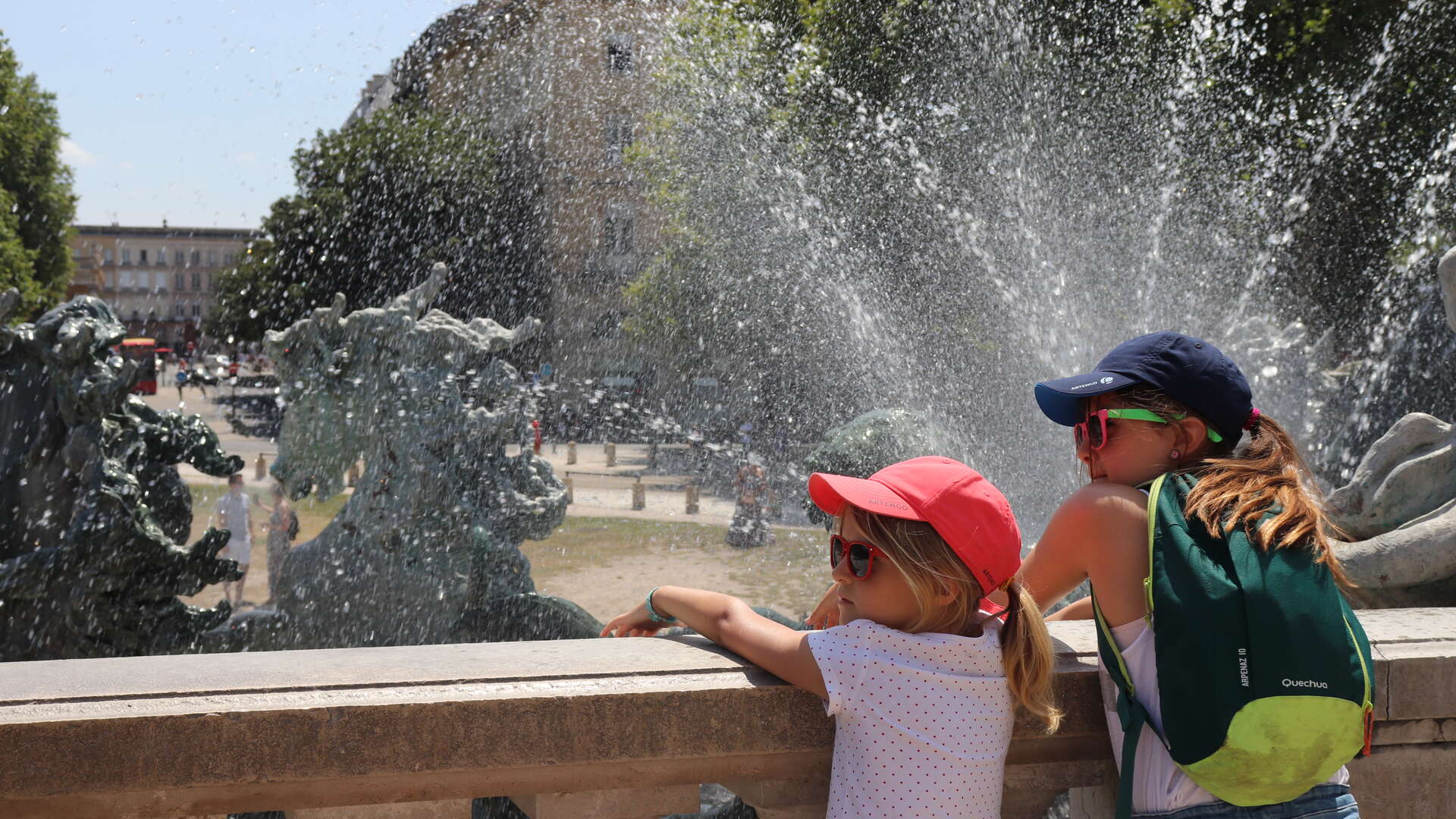 Deux enfants se rafraîchissent dans les jets d'eau d'une fontaine à Bordeaux par une journée ensoleillée, avec la place et ses bâtiments historiques en arrière-plan.
