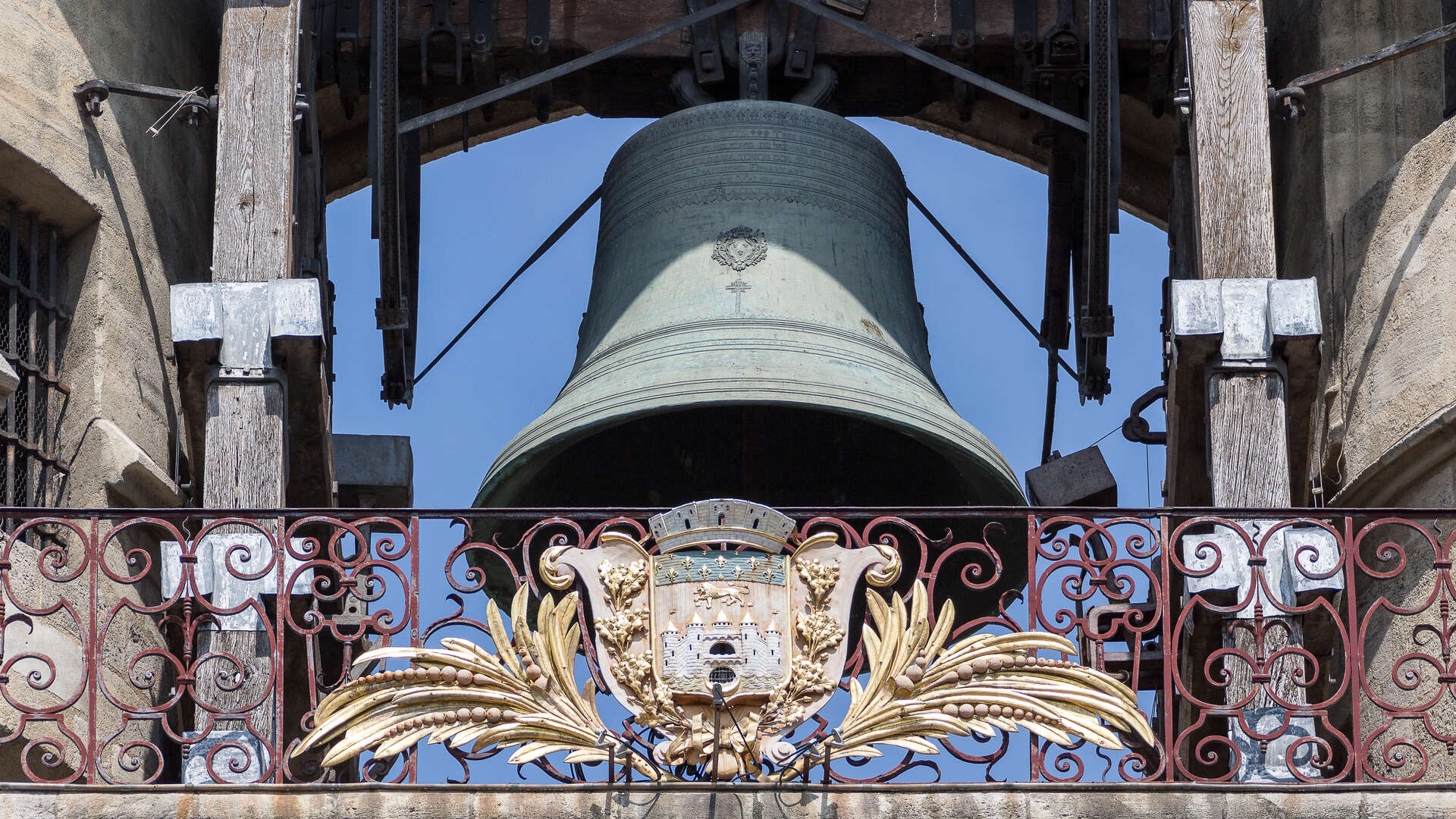La Grosse Cloche de Bordeaux, une imposante cloche en bronze suspendue dans un beffroi en bois, encadrée par une ornementale grille en fer forgé doré aux motifs baroques et un blason sculpté.