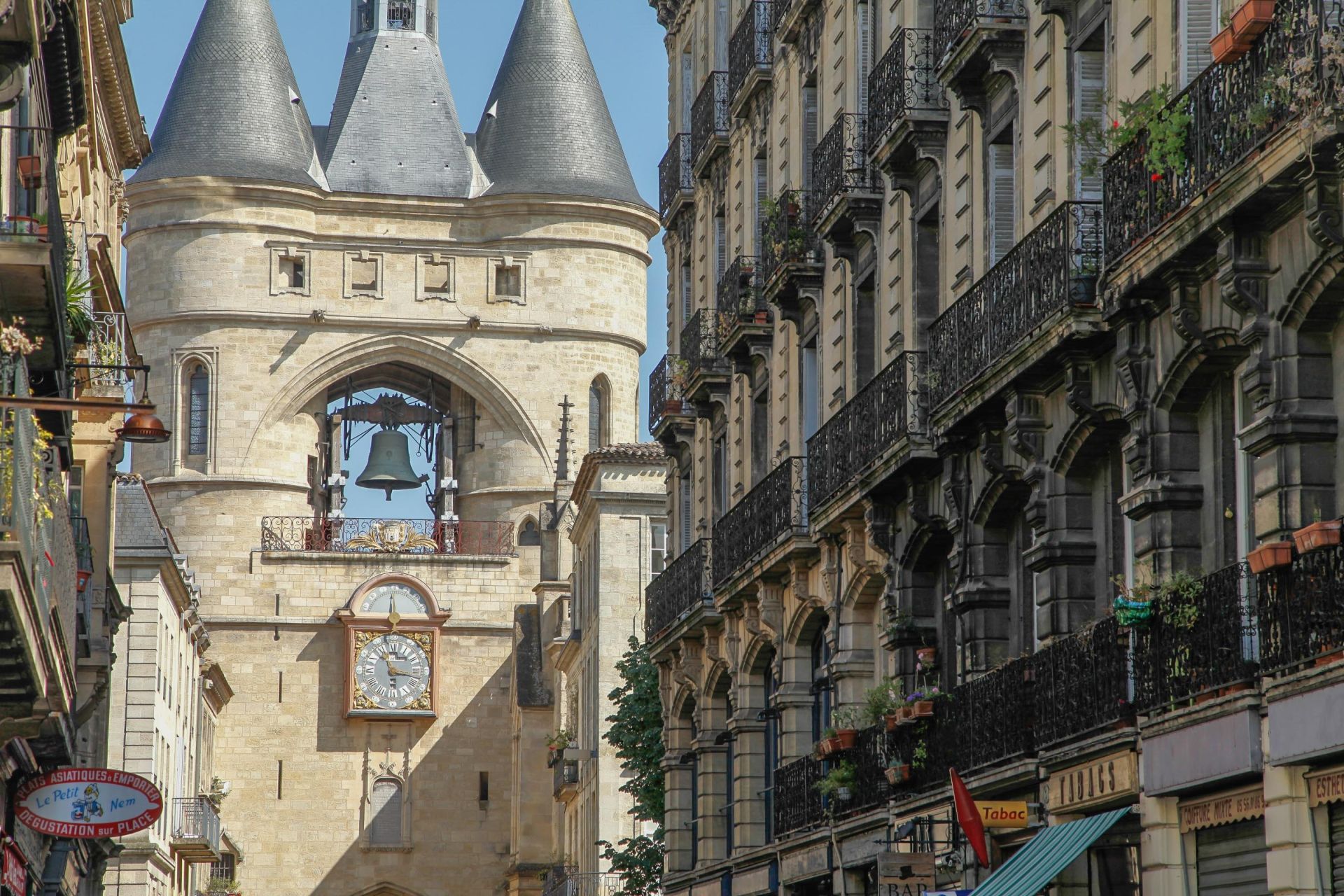 La Porte Cailhau, monument médiéval avec trois tours coniques et une horloge, entourée d'immeubles haussmanniens dans une rue de Bordeaux