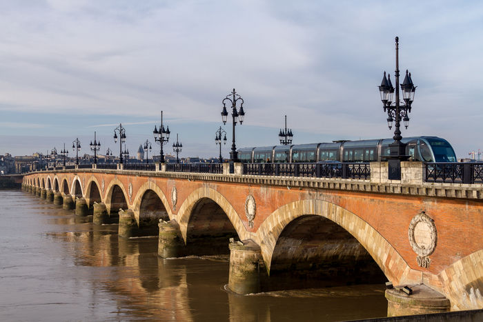 Un train moderne traverse le pont de Pierre de Bordeaux, un pont en brique rouge avec des arches caractéristiques enjambant la Garonne, avec la skyline de la ville en arrière-plan.