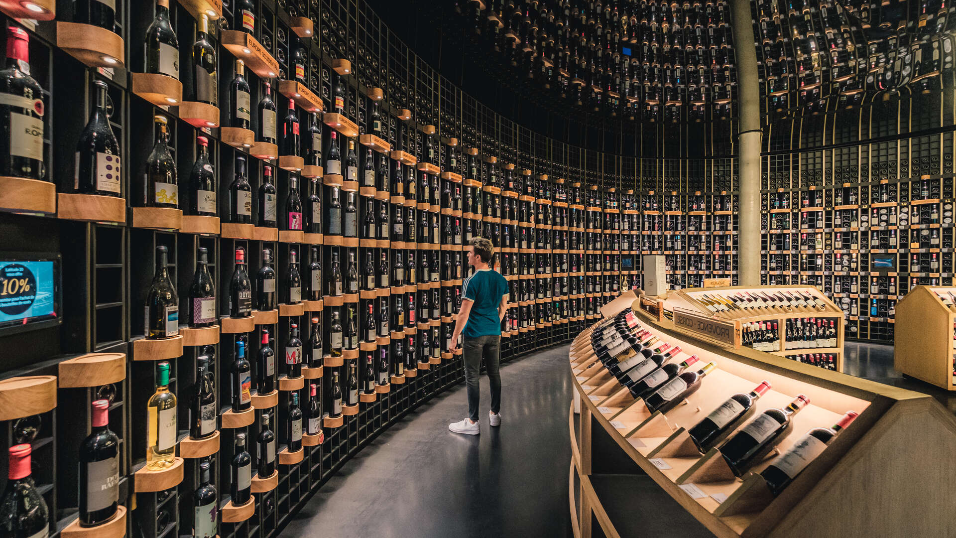 Intérieur d'une cave à vin moderne de Bordeaux avec des murs couverts de bouteilles de vin disposées sur des étagères en bois, un visiteur en tee-shirt bleu observant la collection, et des présentoirs de dégustation au premier plan.