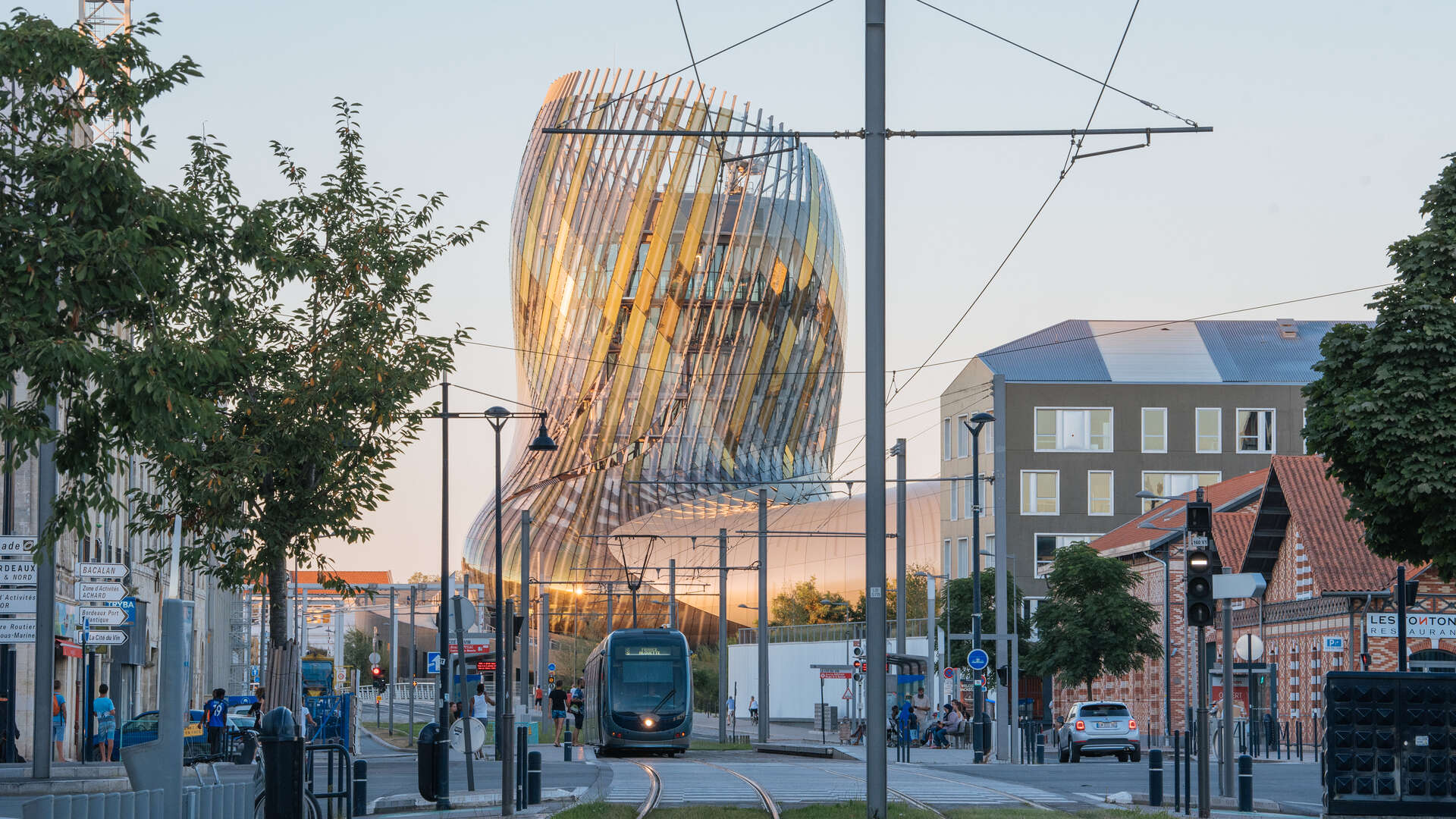 Vue urbaine de Bordeaux avec un tramway moderne passant devant le bâtiment architecturale en verre courbé de la Cité du Vin, entouré de bâtiments historiques et de lignes électriques de tramway.