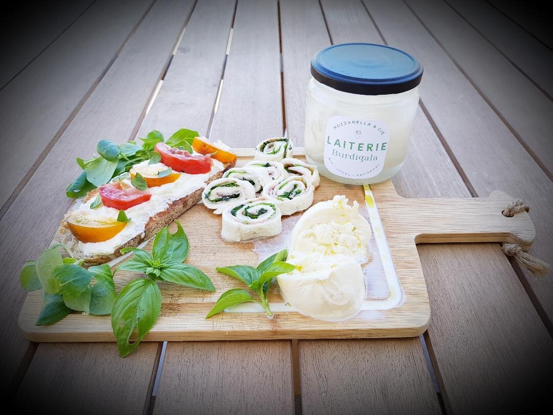 Plateau de dégustation avec pain grillé garni de fromage blanc et légumes frais, rouleaux de mozzarella aux herbes, pot de Laiterie Burdigala, et feuilles de basilic frais sur une planche en bois