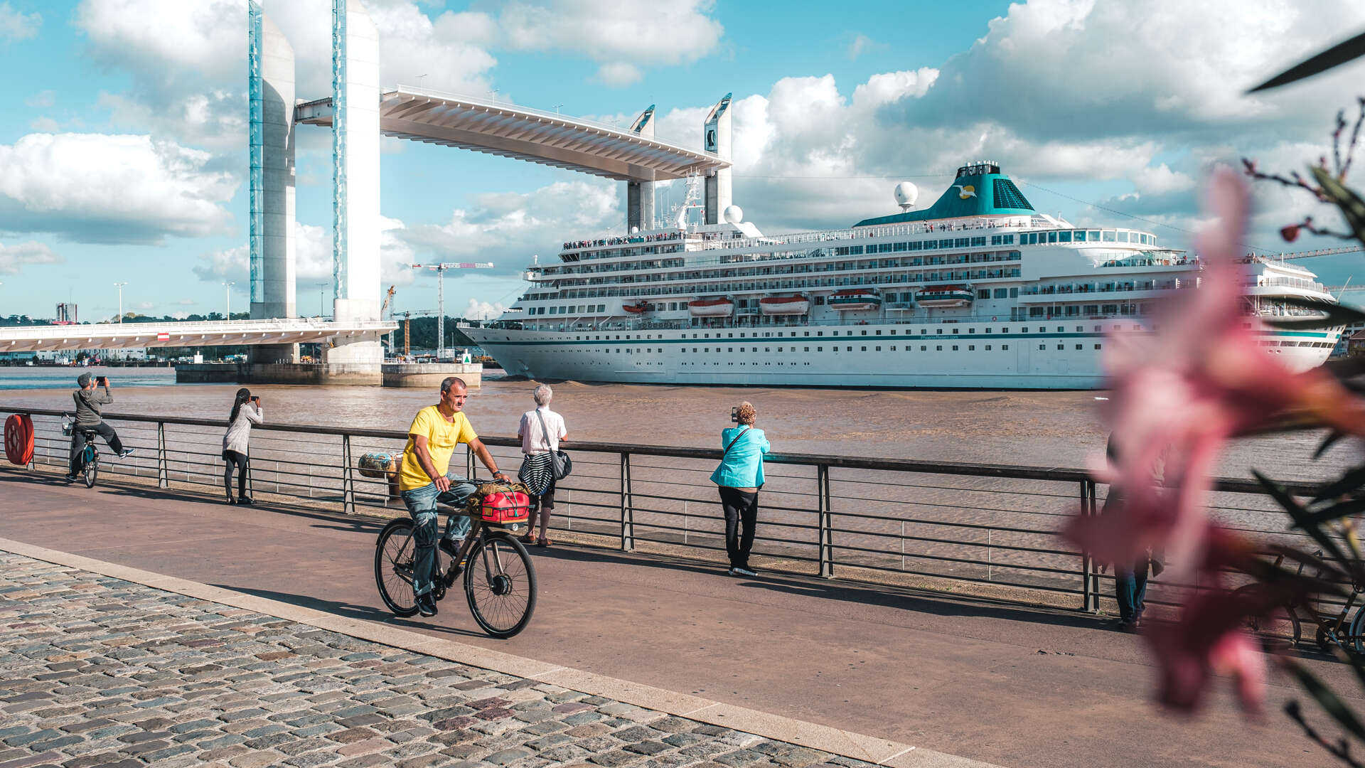 Vue du port de Bordeaux avec un grand paquebot de croisière amarré près du Pont de Pierre, des promeneurs et cyclistes sur la promenade fluviale.