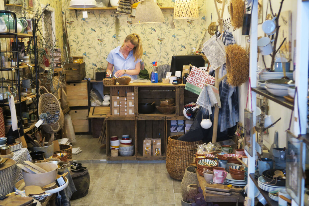 Boutique de décoration intérieure à Bordeaux avec une femme travaillant à son comptoir, entourée de paniers, textiles, objets de décoration et mobilier artisanal.