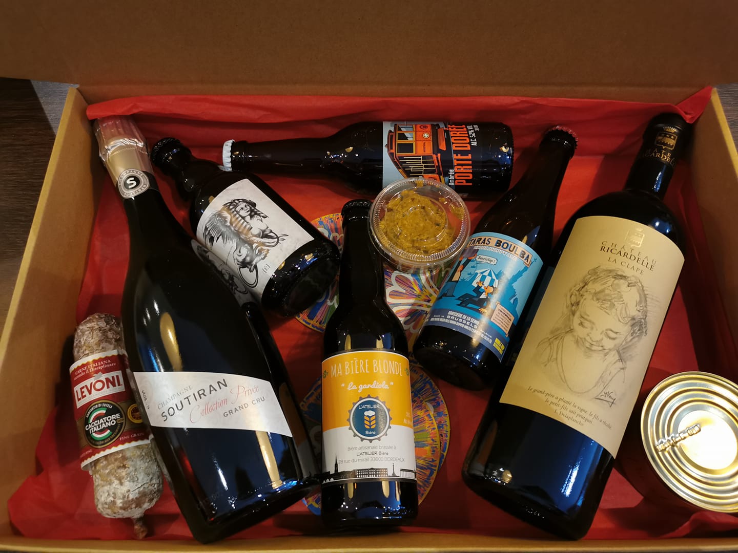 Coffret cadeau de dégustation de vins de Bordeaux contenant plusieurs bouteilles, un saucisson Levoni, des conserves et des accessoires gastronomiques dans une boîte jaune et rouge.