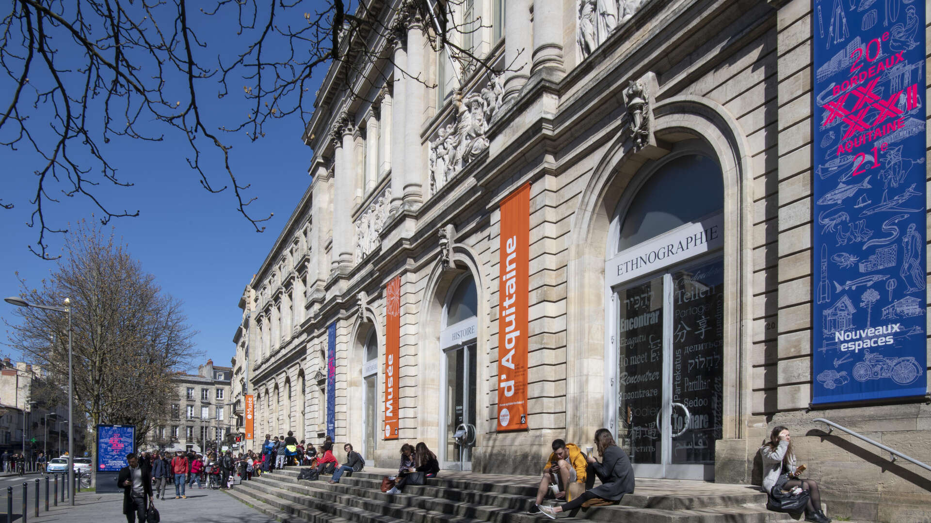 Façade du Musée d'Aquitaine à Bordeaux avec bannières oranges et bleues, visiteurs sur les marches, architecture néoclassique avec colonnes et ornements.