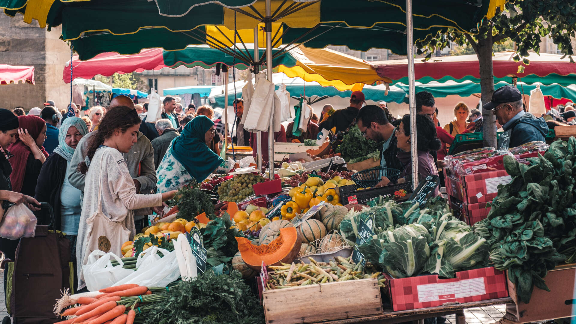 Marché en plein air à Bordeaux avec vendeurs et clients sous des parasols colorés verts, jaunes et rouges, étalant des fruits et légumes frais dont des courges, poivrons, carottes et légumes verts.