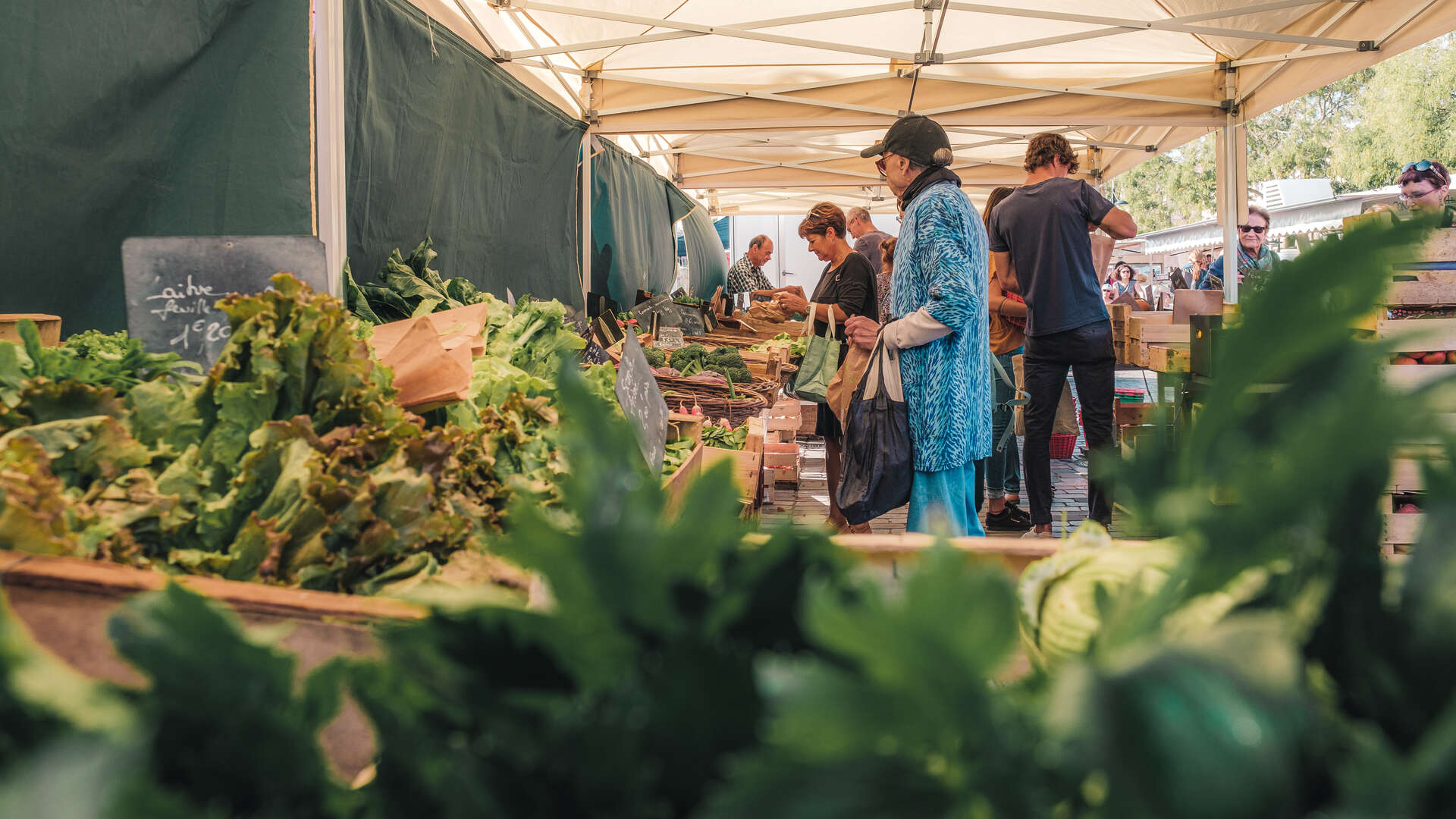 Marché en plein air à Bordeaux sous une tente avec des clients découvrant des légumes frais exposés sur des étals en bois.