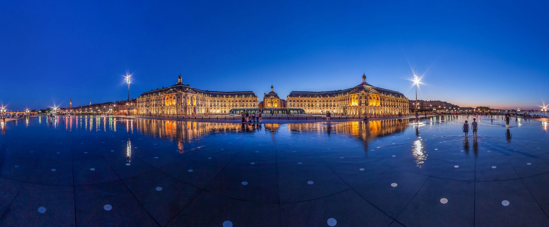 La Place de la Bourse de Bordeaux illuminée à la nuit tombante, avec ses bâtiments classiques se reflétant dans le miroir d'eau, un tramway bleu passant devant, et des personnes marchant sur la surface mouillée.