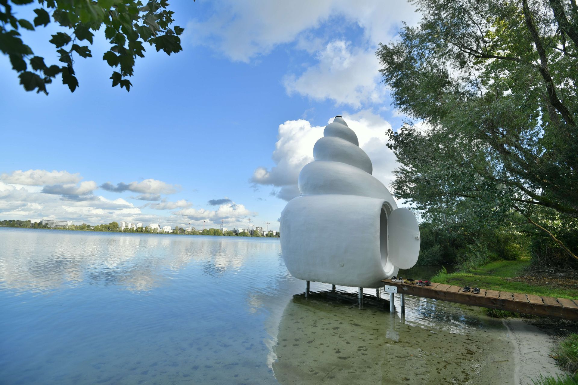 Installation artistique blanche en forme de coquillage au bord de l'eau à Bordeaux, avec la ville en arrière-plan et des arbres verts.