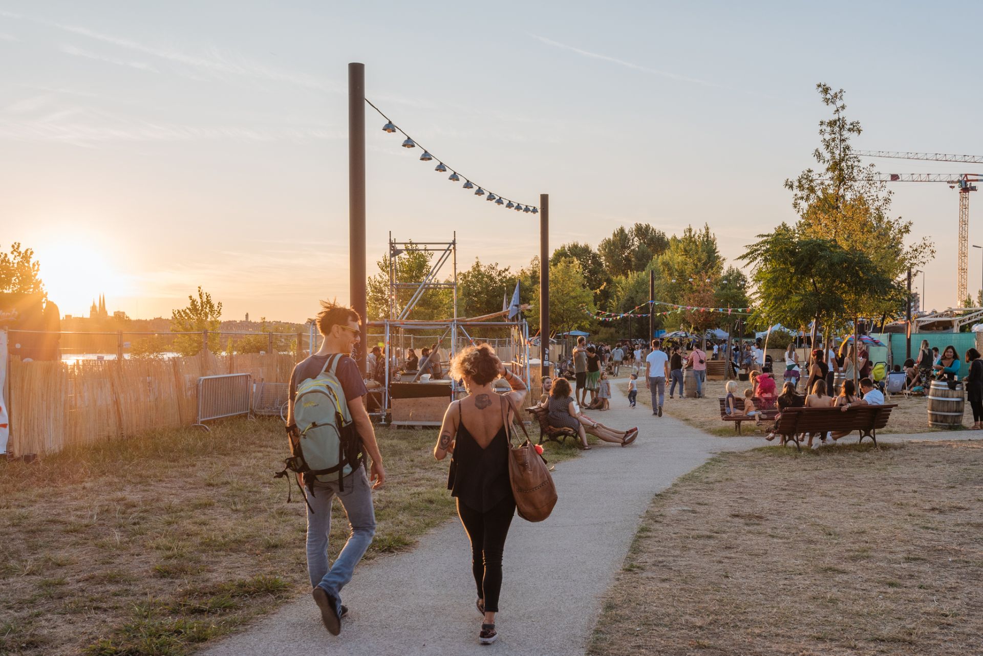 Deux jeunes femmes courent sur un sentier dans un parc urbain au coucher du soleil, avec des installations de jeux et des visiteurs en arrière-plan au Parc aux Angéliques de Bordeaux.