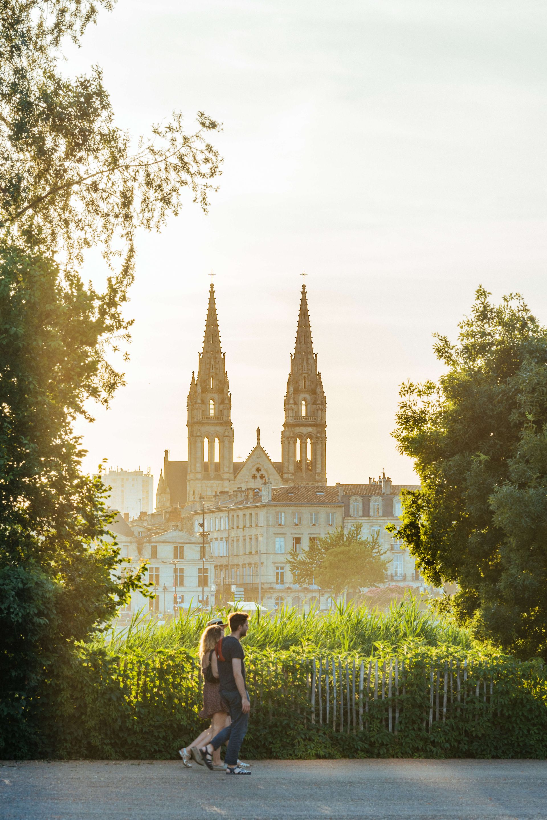 Deux personnes marchent sur un chemin bordé d'arbres avec la Basilique Saint-Michel de Bordeaux visible en arrière-plan au coucher du soleil.