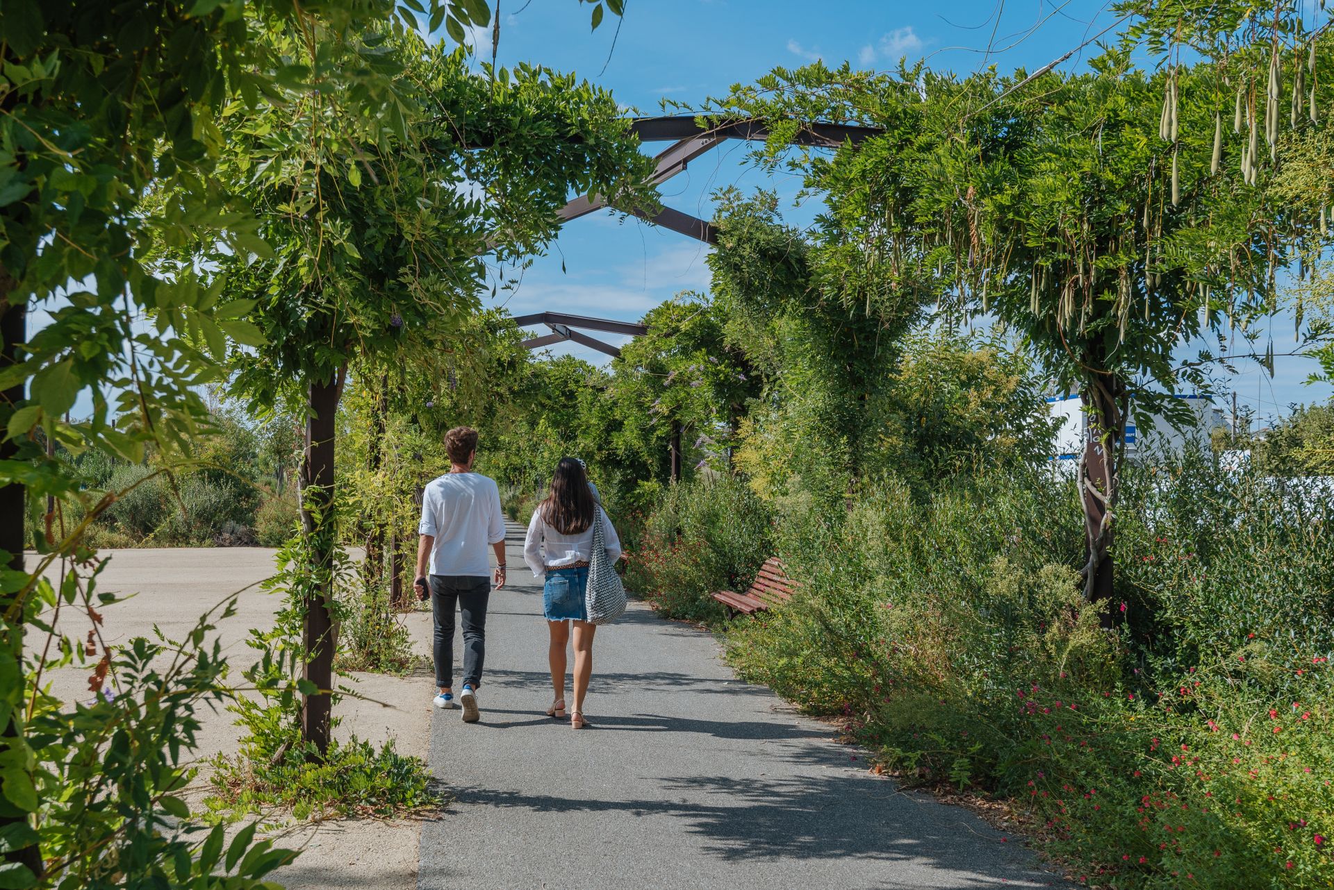 Deux personnes marchent sur un sentier verdoyant du Parc aux Angéliques à Bordeaux, encadrées par une végétation luxuriante et des structures métalliques arquées.