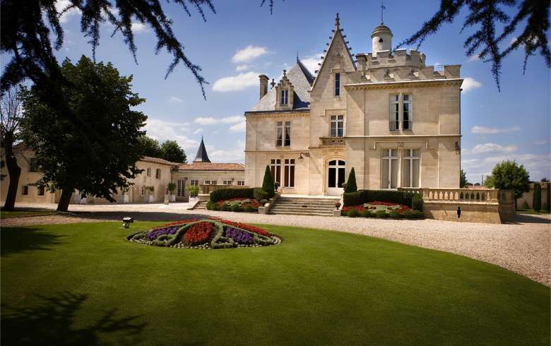 Château de style Belle Époque avec tours pointues et façade en pierre beige, entouré d'un parc paysager avec pelouse verte et parterre de fleurs colorées, situé dans la région de Bordeaux.