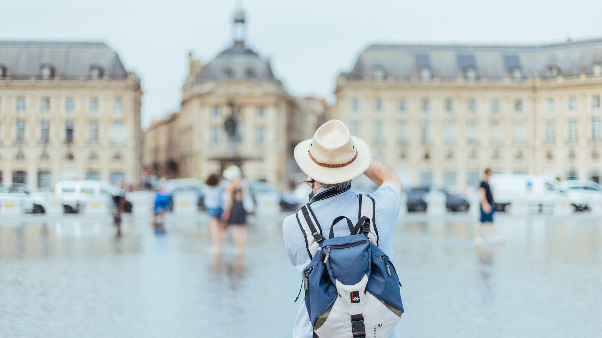 Touriste de dos portant un chapeau blanc et un sac à dos bleu, contemplant la Place de la Bourse et ses bâtiments historiques à Bordeaux