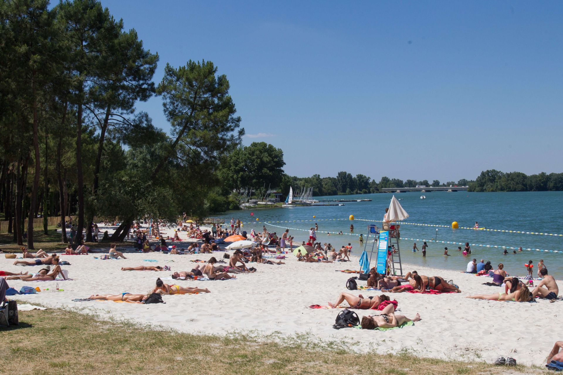 Plage aménagée au bord d'un lac avec de nombreux baigneurs, des voiliers, une zone de baignade délimitée et des arbres en arrière-plan sous un ciel dégagé.