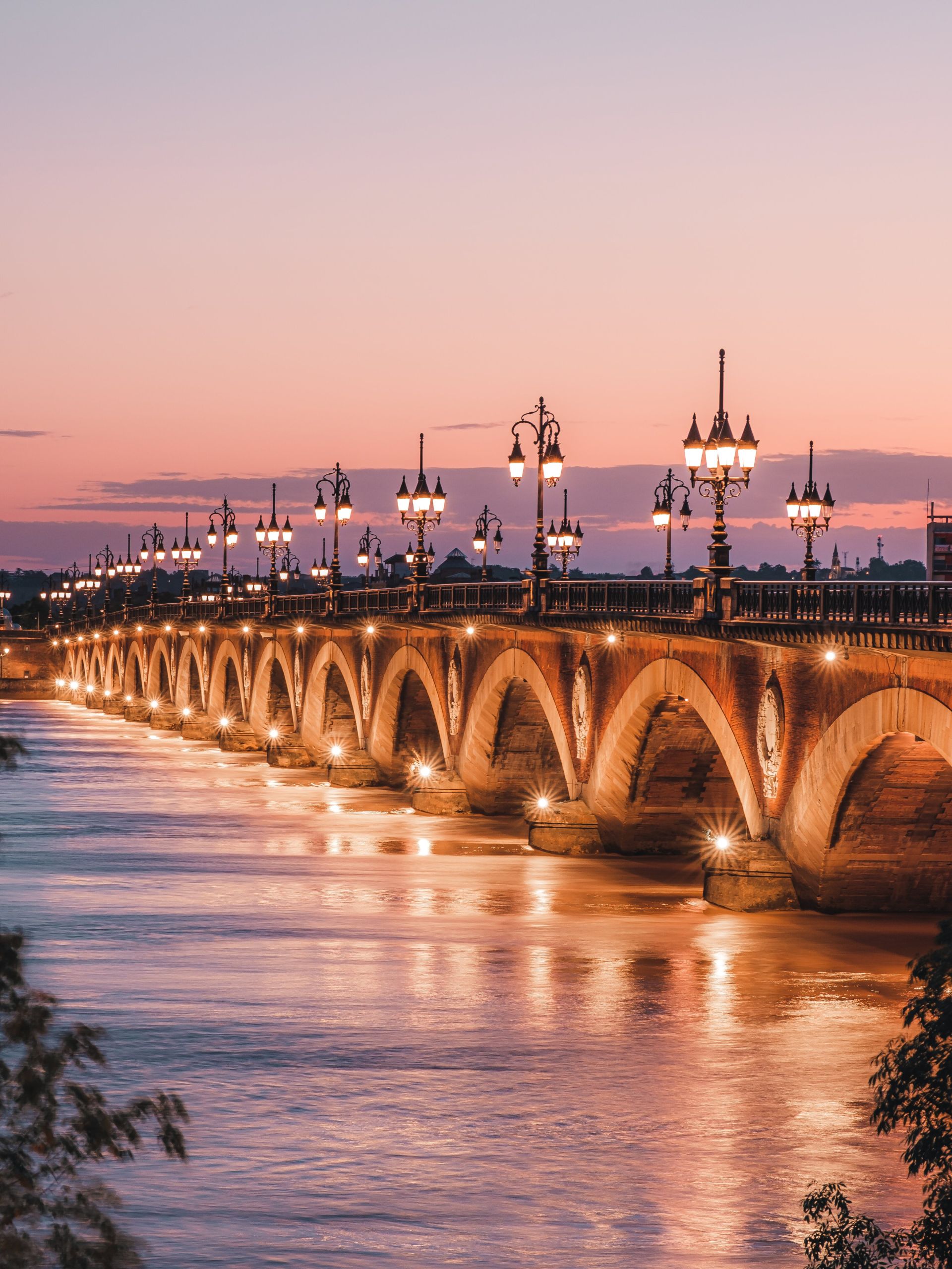 Le Pont de Pierre de Bordeaux illuminé au coucher du soleil, avec ses arches en pierre et ses réverbères allumés se reflétant dans l'eau de la Garonne