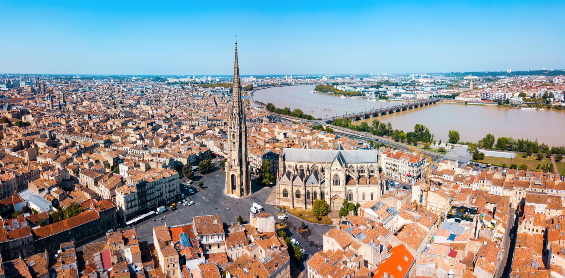 Vue aérienne de Bordeaux montrant la Basilique Saint-Michel avec sa flèche caractéristique, la Garonne avec ses ponts, et l'urbanisation dense de la ville historique aux toits orangés.