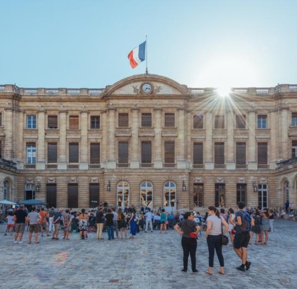 La Cour du Parlement de Bordeaux avec sa façade classique en pierre, le drapeau français flottant au-dessus, et des visiteurs rassemblés sur la place pavée sous un ciel dégagé.