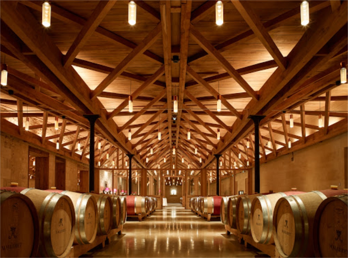 Intérieur d'un chai de Bordeaux avec des rangées de fûts de chêne alignés symétriquement sous une structure architecturale en bois aux poutres apparentes, éclairé par des luminaires modernes.