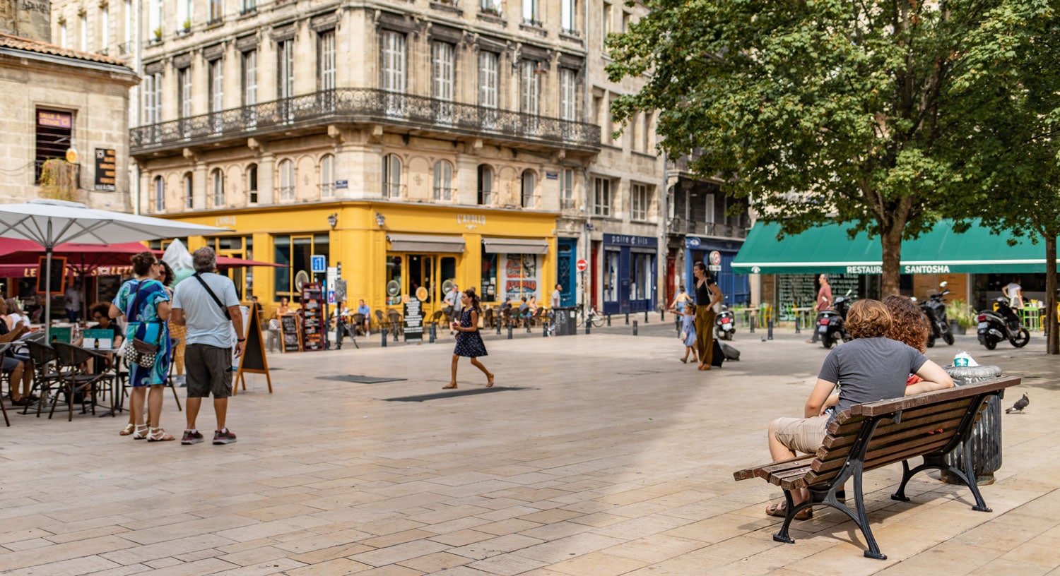 Une place publique à Bordeaux avec des bâtiments haussmanniens, un commerce jaune vif au rez-de-chaussée, des terrasses de café avec parasols, des arbres verts, et des passants se détendant sur des bancs.