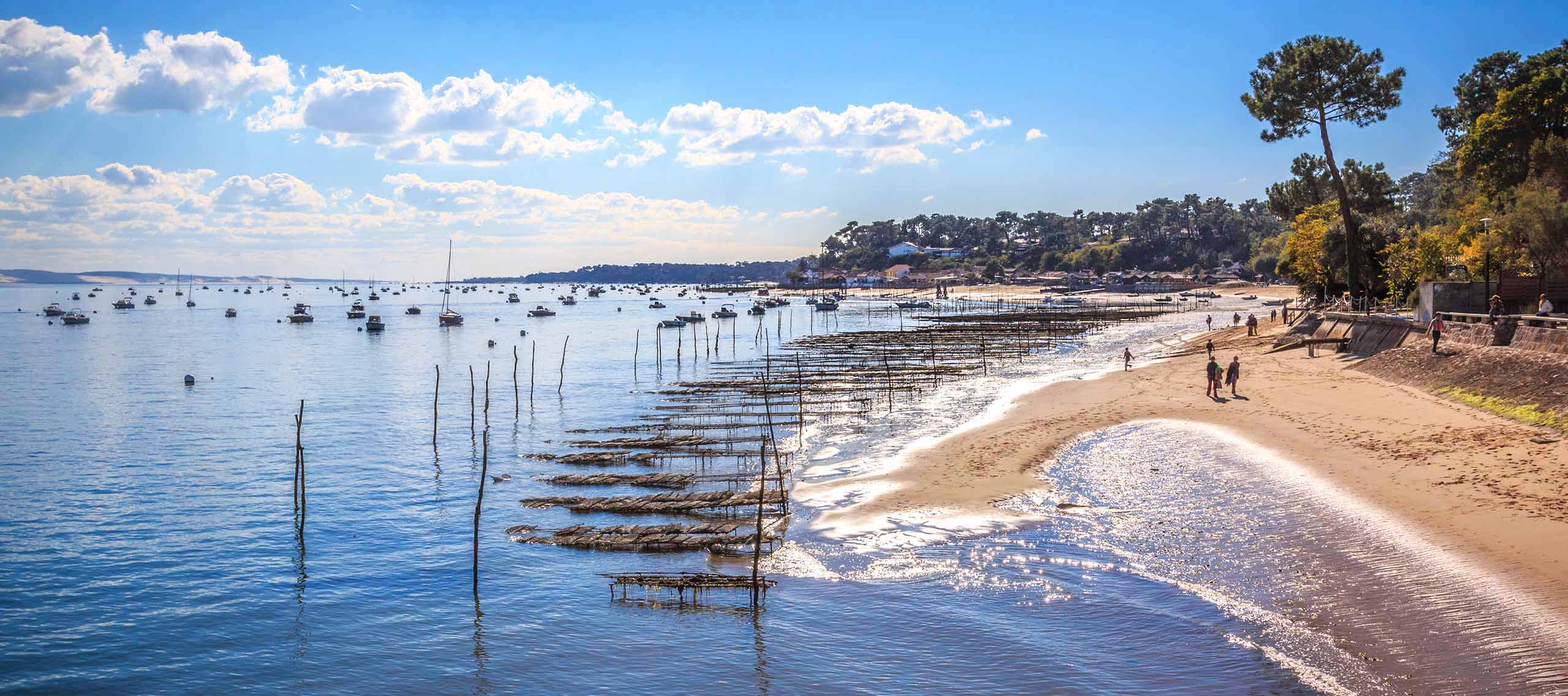 Plage avec sable fin, voiliers ancrés dans une baie calme, parcs à huîtres, végétation côtière et personnes se baignant sous un ciel bleu avec nuages blancs