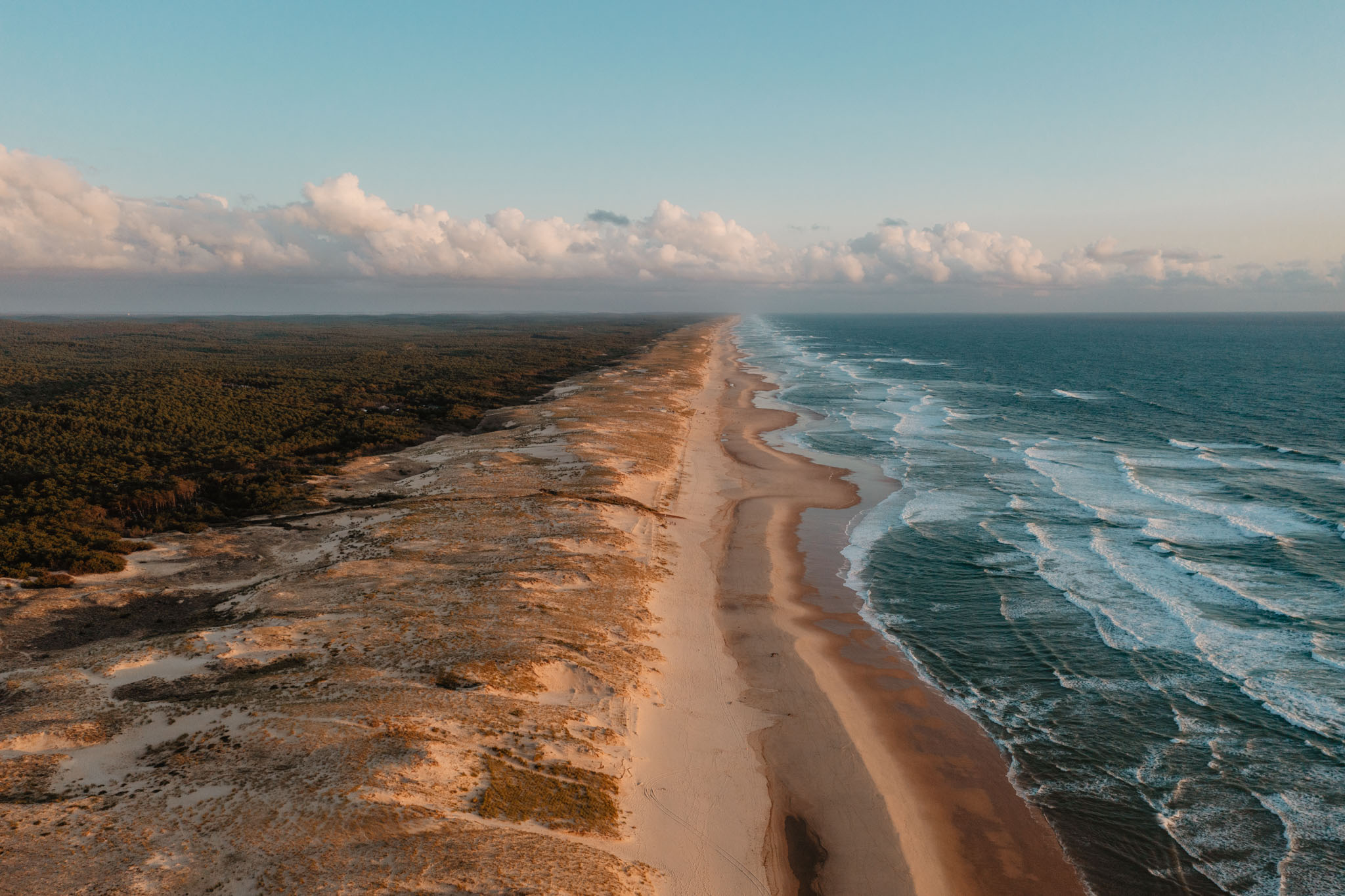 Vue aérienne d'une longue plage de sable bordée par l'océan avec des vagues, une forêt dense à gauche et un ciel nuageux