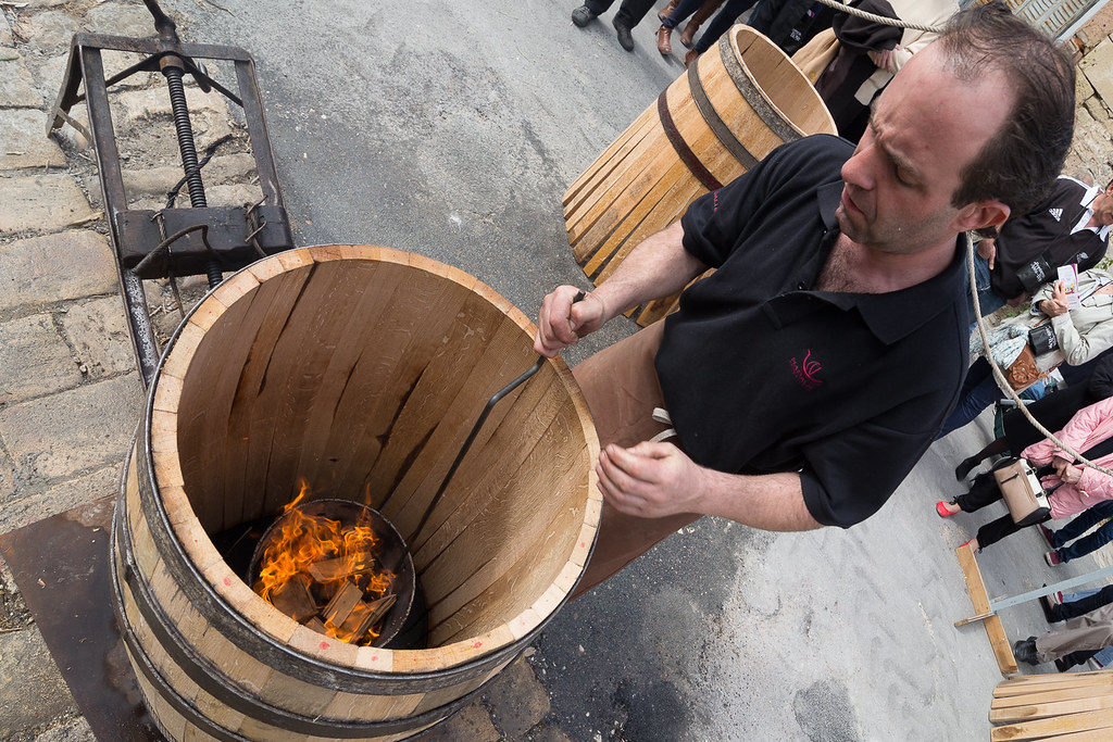 Un homme en polo noir tient un grand tonneau en bois avec un feu ardent à l'intérieur, lors d'une démonstration traditionnelle à Bordeaux.