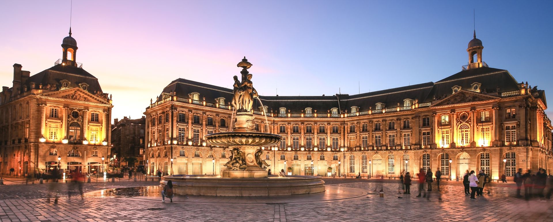 Place de la Bourse à Bordeaux illuminée la nuit, avec ses bâtiments historiques en pierre de taille, une fontaine centrale et des personnes se promenant sur la place pavée.